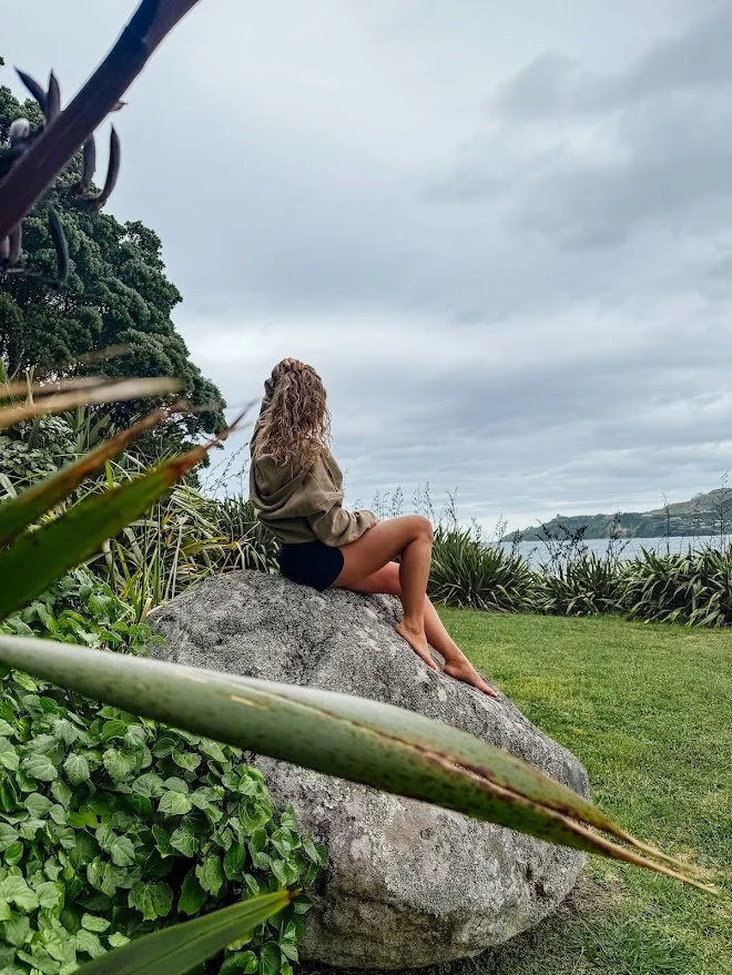 Person sitting on a large rock surrounded by greenery, overlooking a body of water under a cloudy sky.