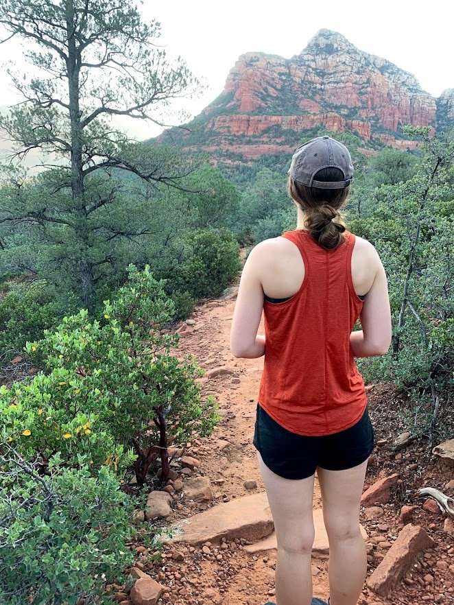 A woman in a red tank top, black shorts, and gray baseball cap hiking on a dirt trail through green bushes and trees towards a red rock mountain in the background.