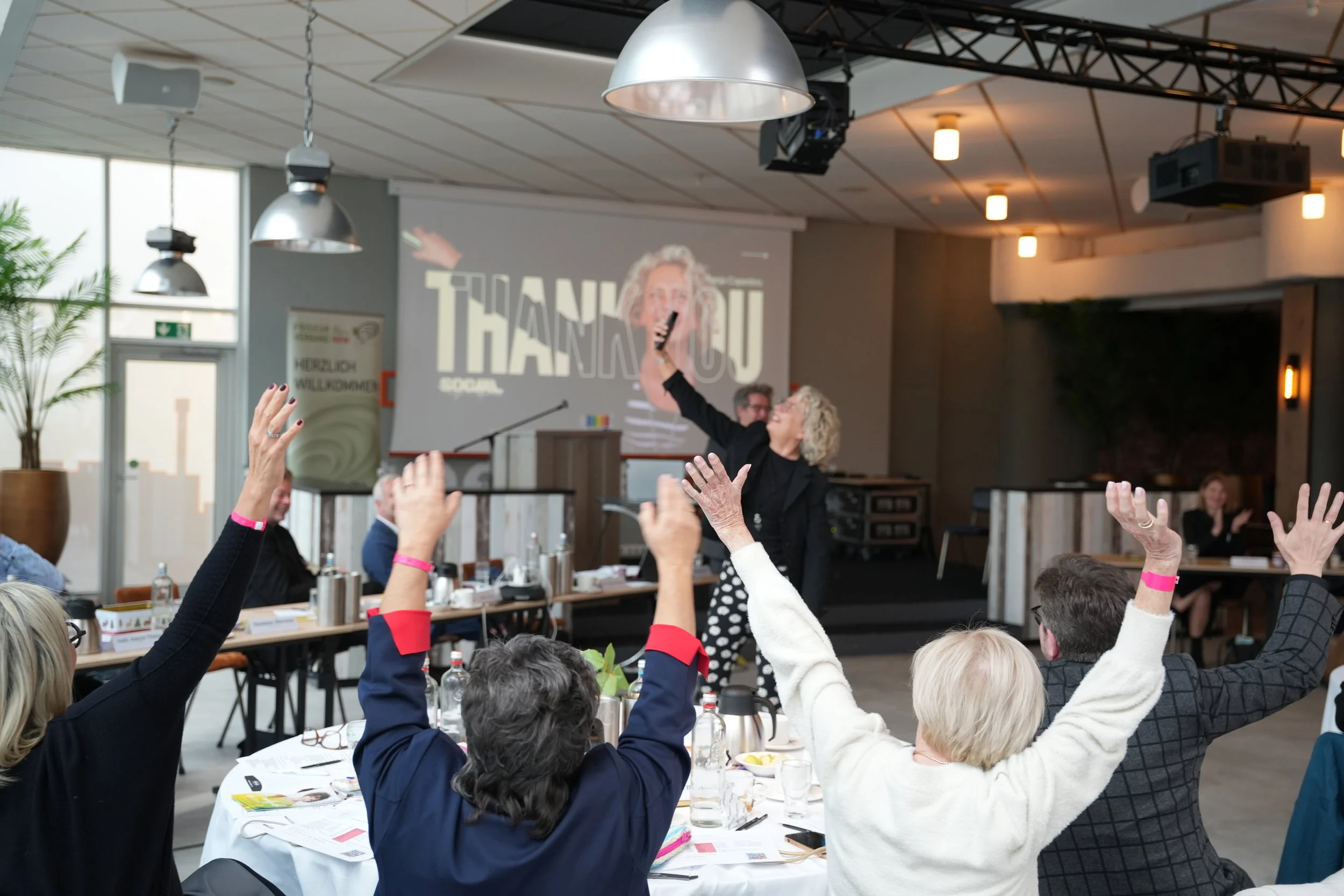 Menschen während einer Tagung vom LIV NRW in Kalkar, die Friseure heben die Hände, im Hintergrund ist Tanja Copertino auf der Bühne, die eine Präsentation über Instagram hält, auf einer Leinwand steht 'Thank You'.