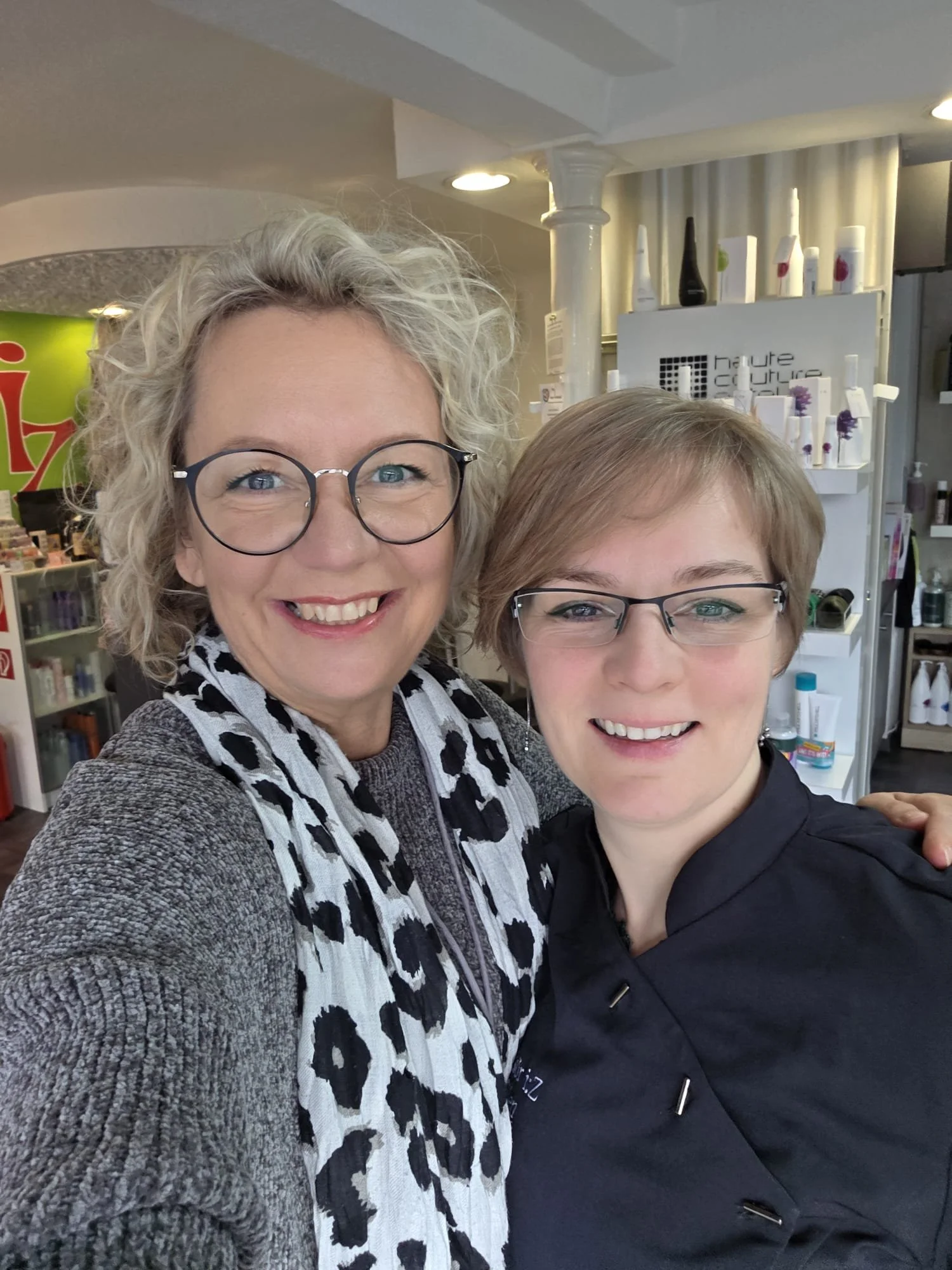 Tanja Copertino macht mit einer Kundin ein Foto, die sich in einem Friseur befinden. Beide Lachen freundlich in die Kamera.