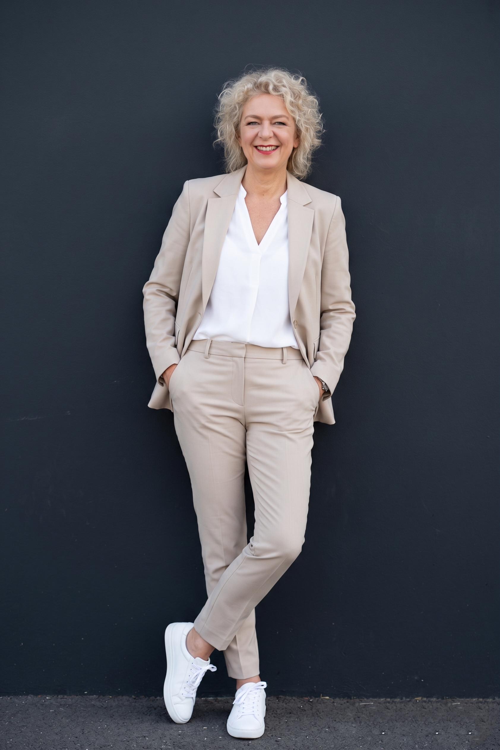 Tanja Copertino mit lockigem, blonden Haar trägt einen beigen Anzug, weiße Schuhe und steht vor einer dunklen Wand. Sie lächelt, ihre Hände sind in den Taschen, und sie trägt einen weißen Hemd unterm Blazer.