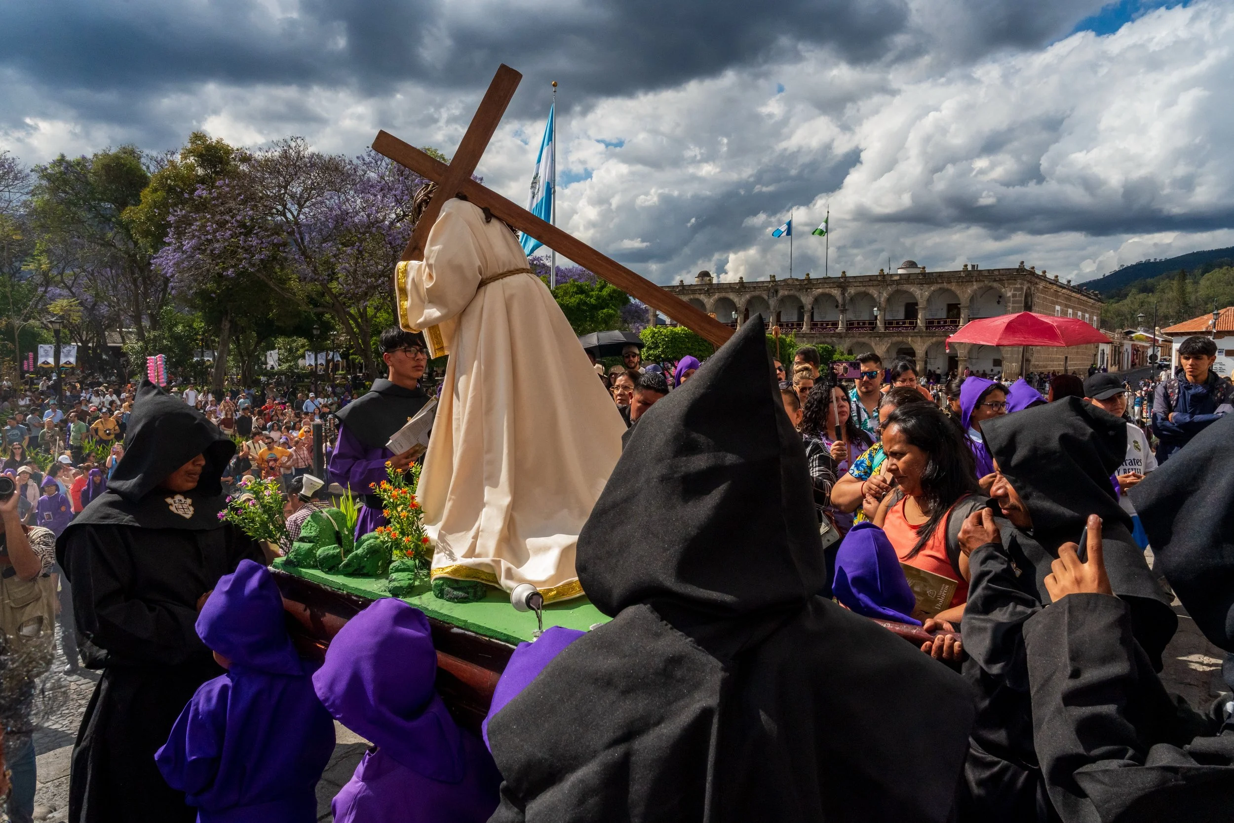 Jésus en tunique crème avec la croix, cagoule noire au premier plan, arcades et drapeaux derrière