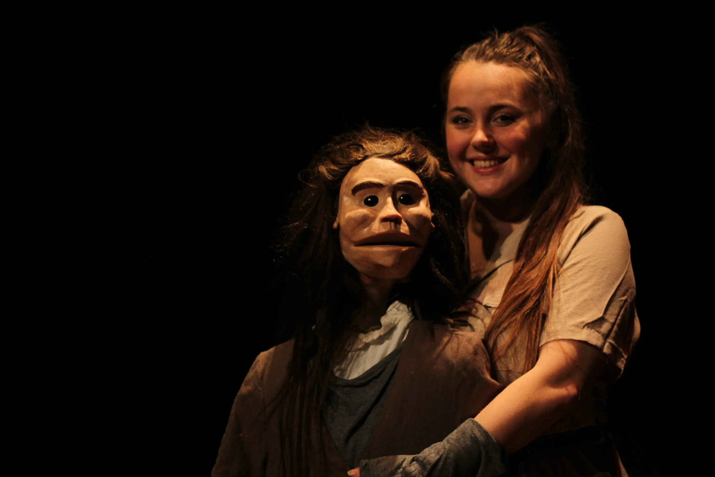 Willemijn Hordijk die glimlacht en een pop vasthoudt met een menselijk gezicht en lang haar op een donker podium.