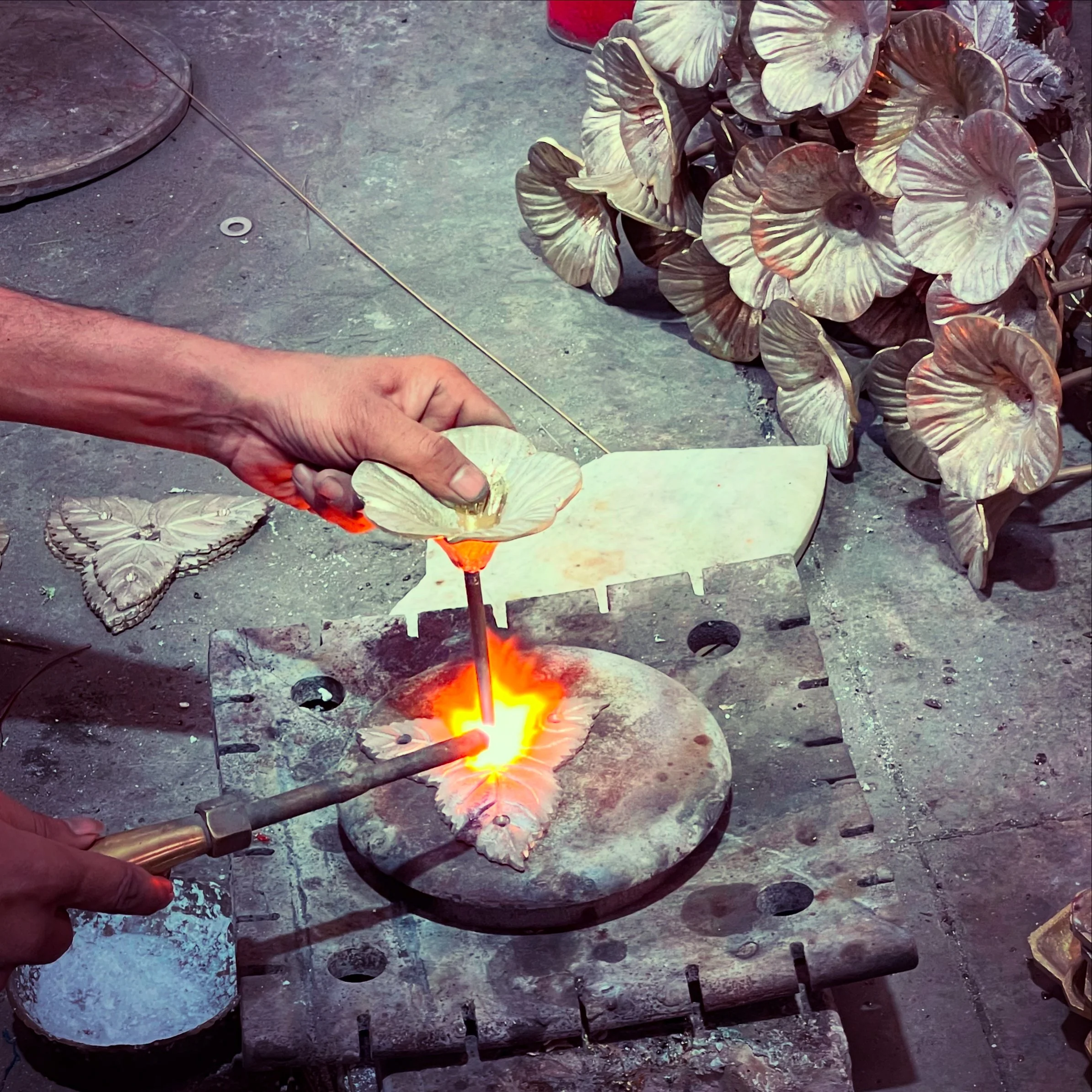 A person holding a glowing hot metal piece with tongs over a forge with a fire, surrounded by metal tools and a work surface.