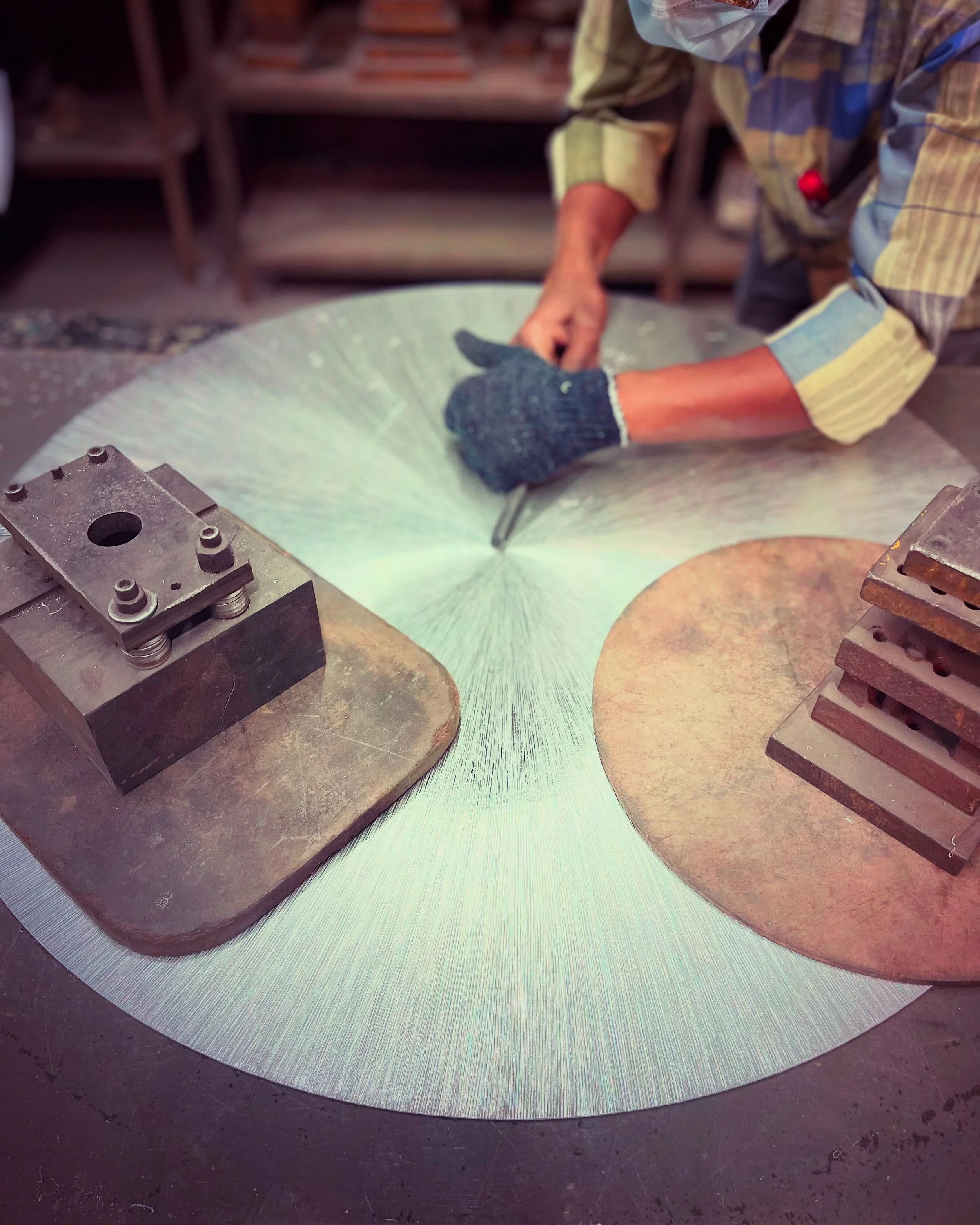 A person working on a large circular metal workpiece in a workshop, using a hand tool, with the workpiece secured to a workbench with metal clamps.