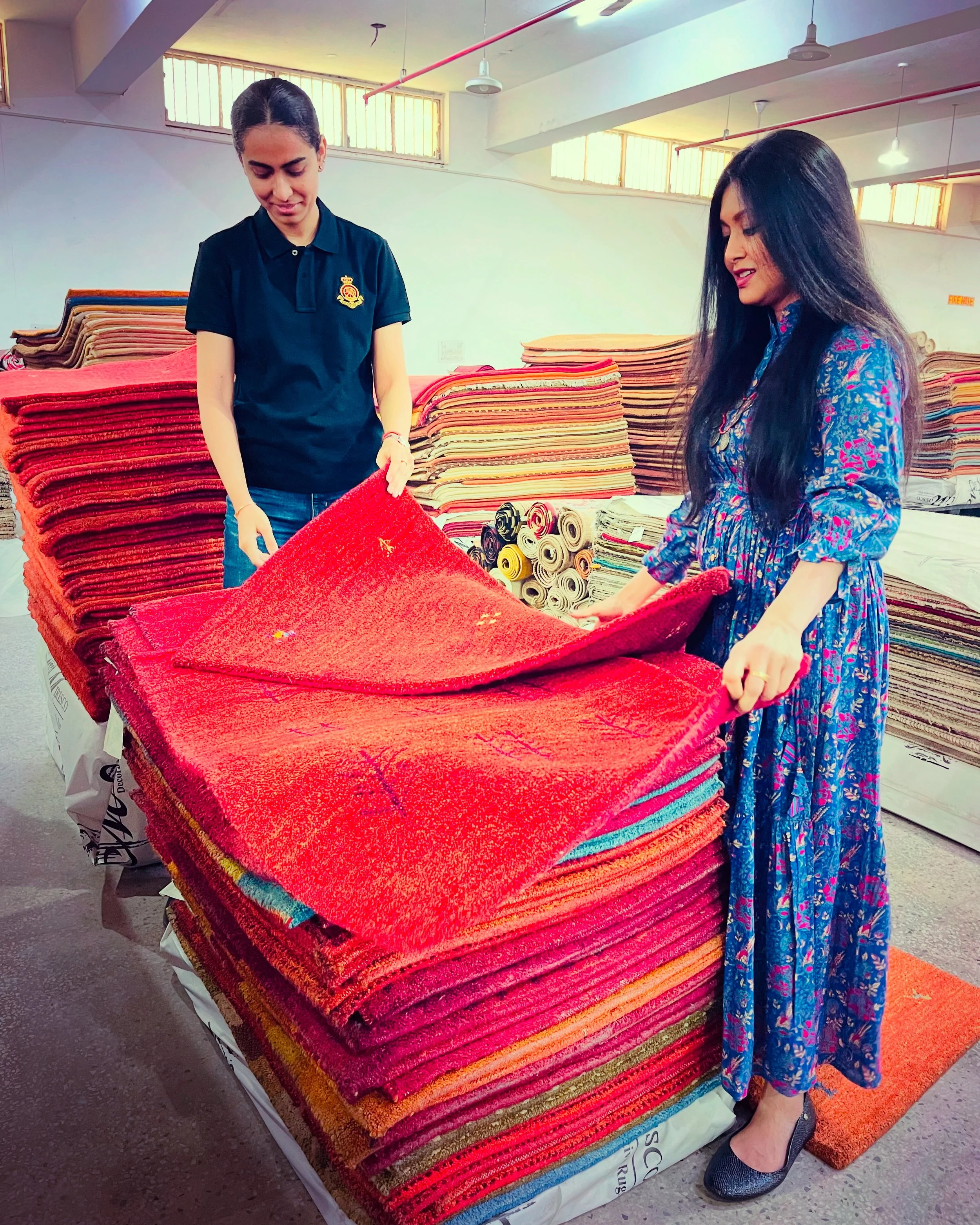 Two women shopping for carpets in a store, examining a red rug. One woman, wearing a black uniform with a crest on the chest, holds the rug while the other woman, dressed in a blue floral dress, looks on. The store has other colorful rugs stacked on tables in the background, with sunlight coming through windows.
