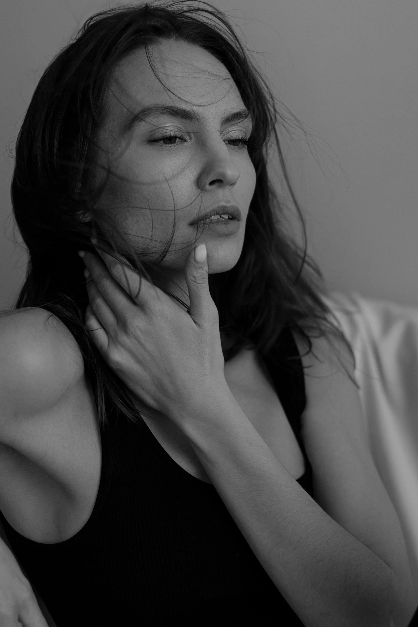 Black and white close-up photograph of a woman with long hair, her eyes partially closed, touching her neck with one hand, wearing a sleeveless top.