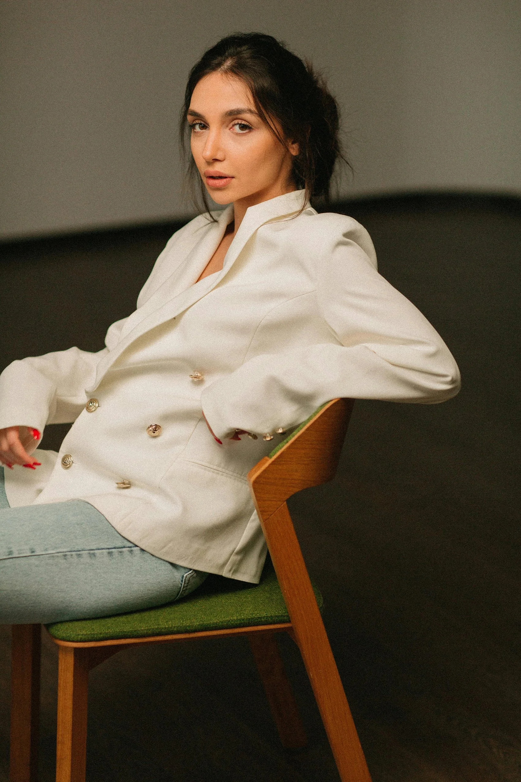 A woman with dark hair, dressed in a white blazer and light jeans, sitting on a green-cushioned wooden chair against a dark background.