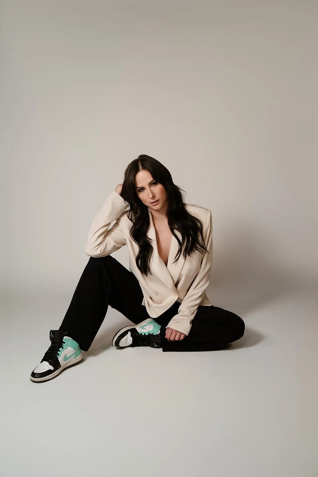 A woman with dark hair sitting on the floor with one leg crossed over the other, wearing a beige blazer, black pants, and sneakers with turquoise accents, posing against a plain light-colored background.
