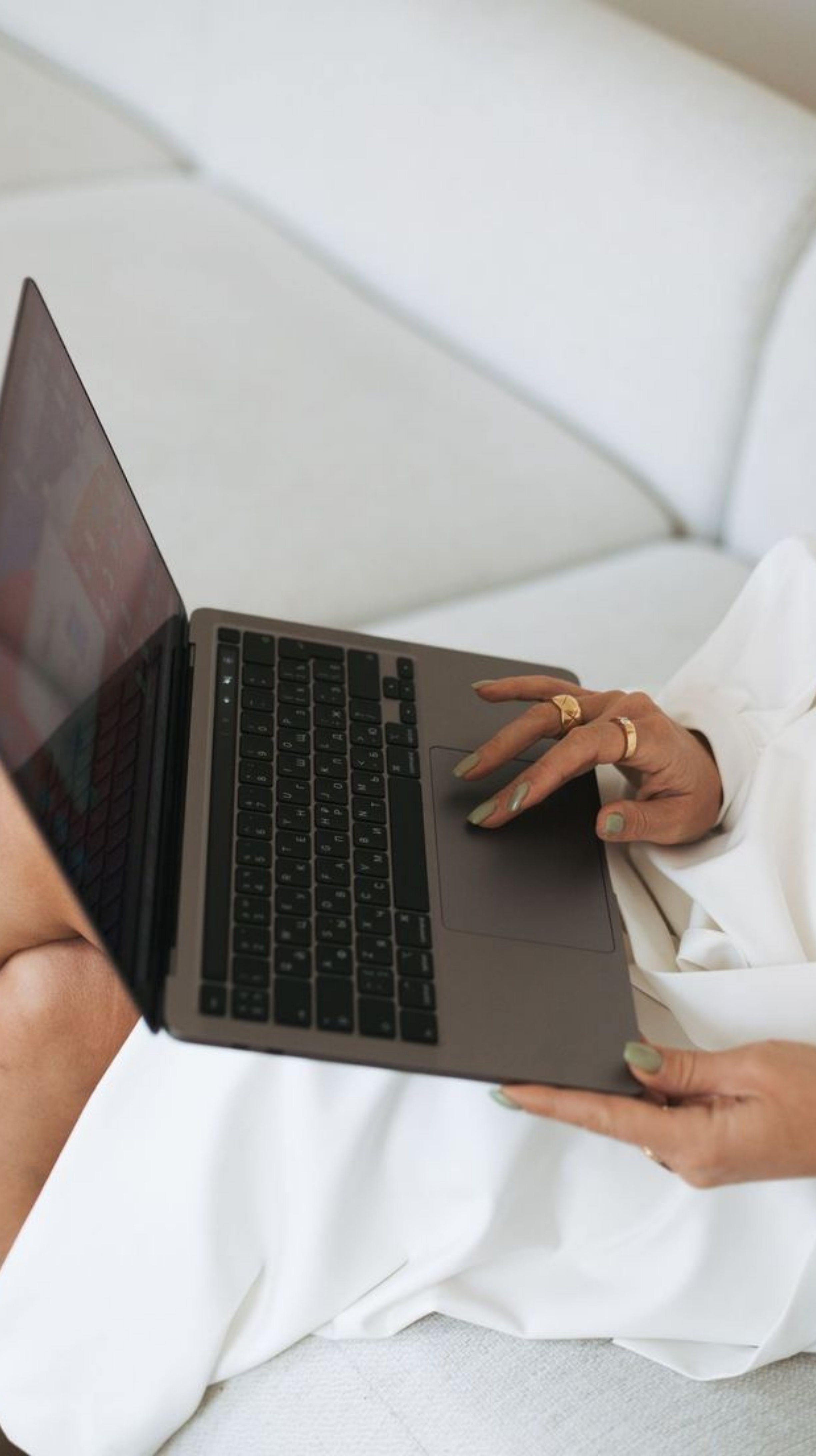 A person with painted nails and multiple rings uses a laptop on their lap, sitting on a white couch.