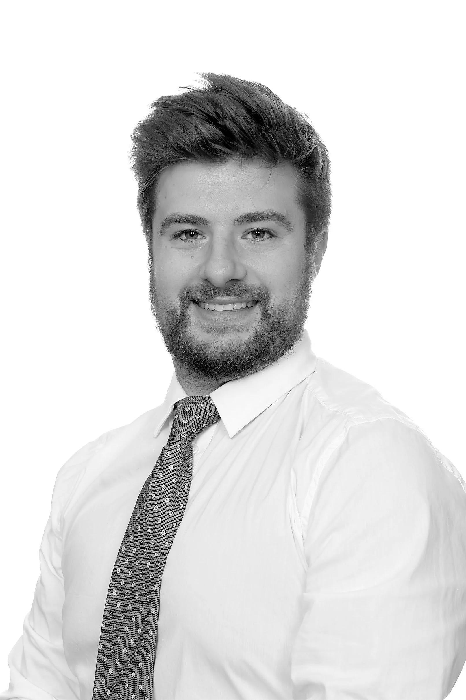 Black-and-white portrait of a smiling man with a beard, wearing a white shirt and a patterned tie, against a plain white background.
