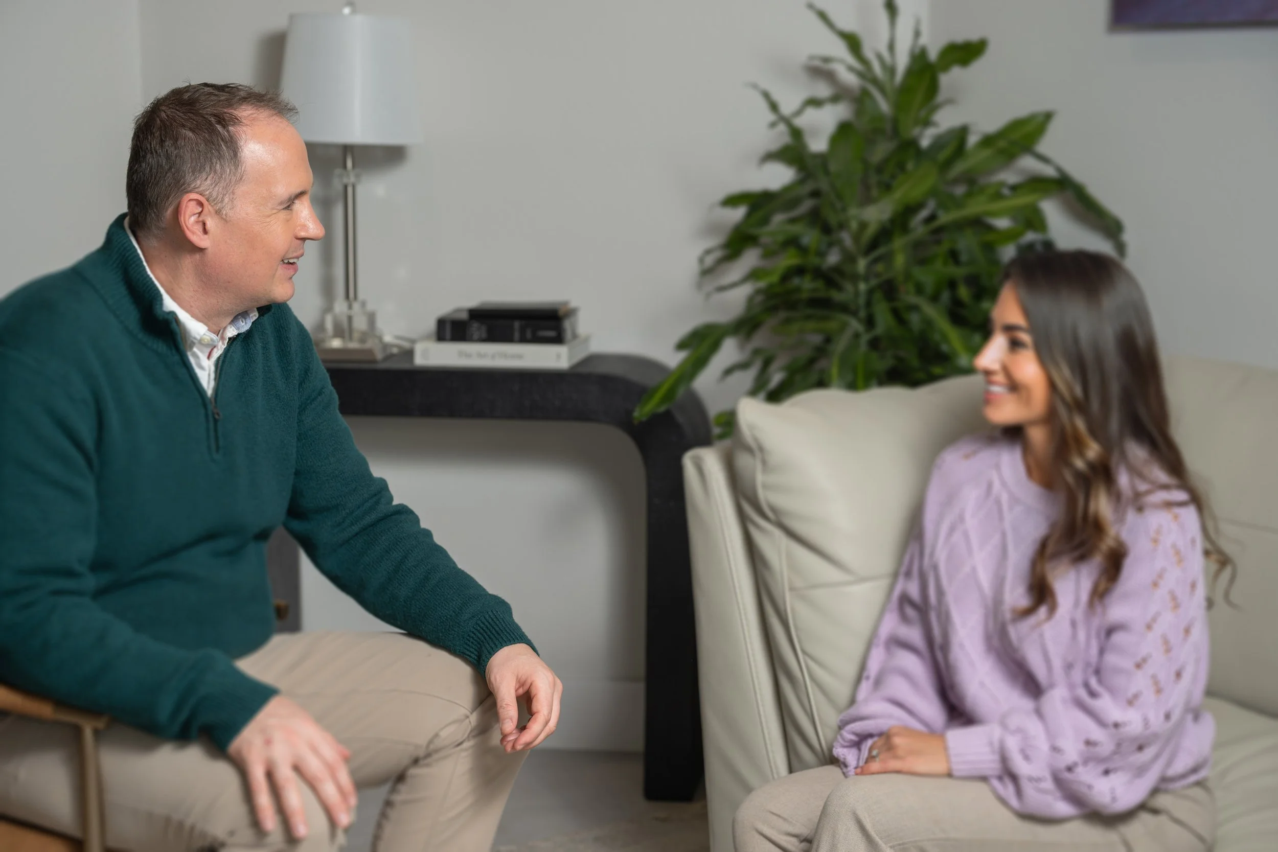 A man in a navy hoodie sitting and talking in a therapy session, with a glass of water and tissue box on a side table nearby.