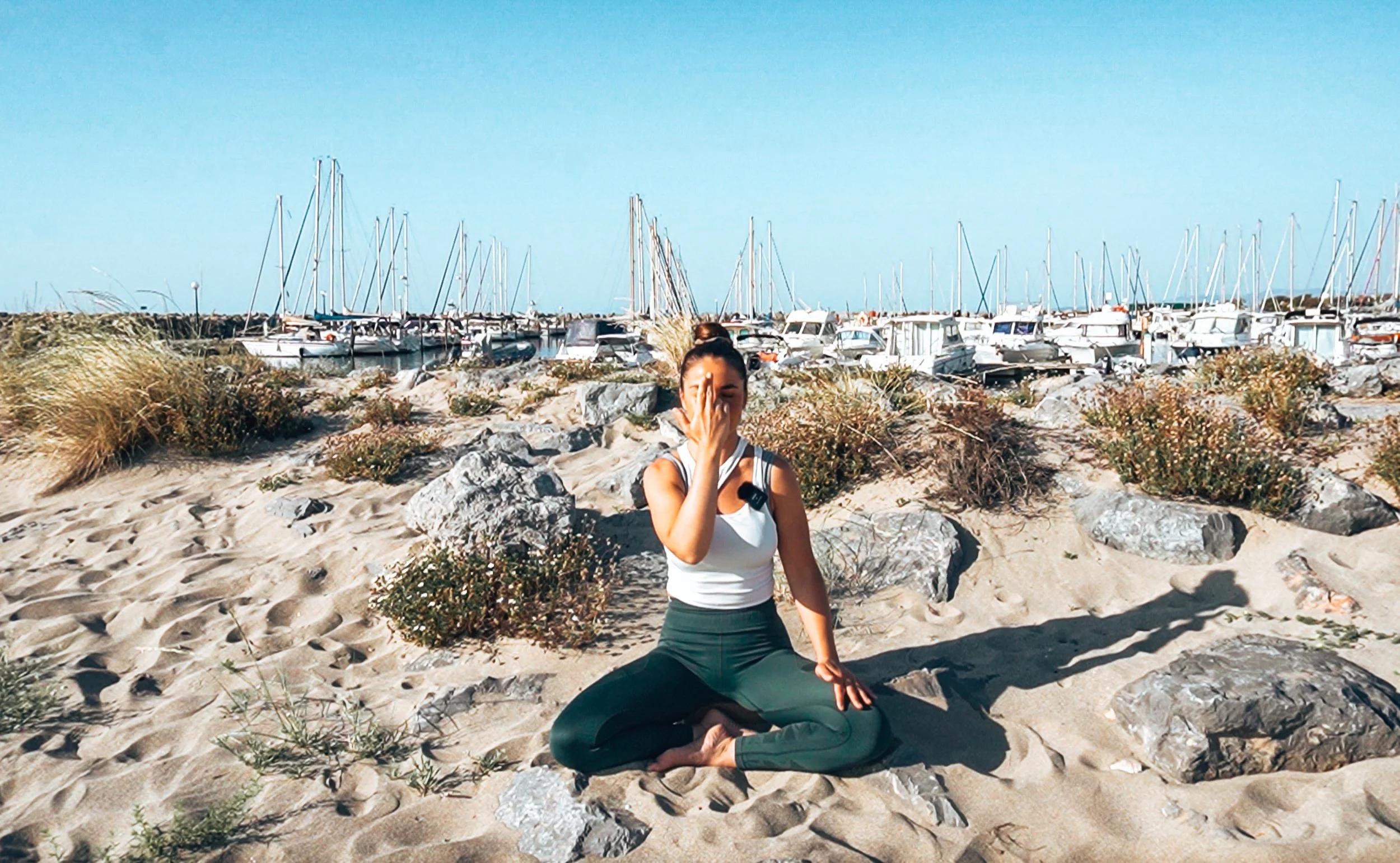 Femme méditant en tailleur sur la plage avec des bateaux à voile en arrière-plan