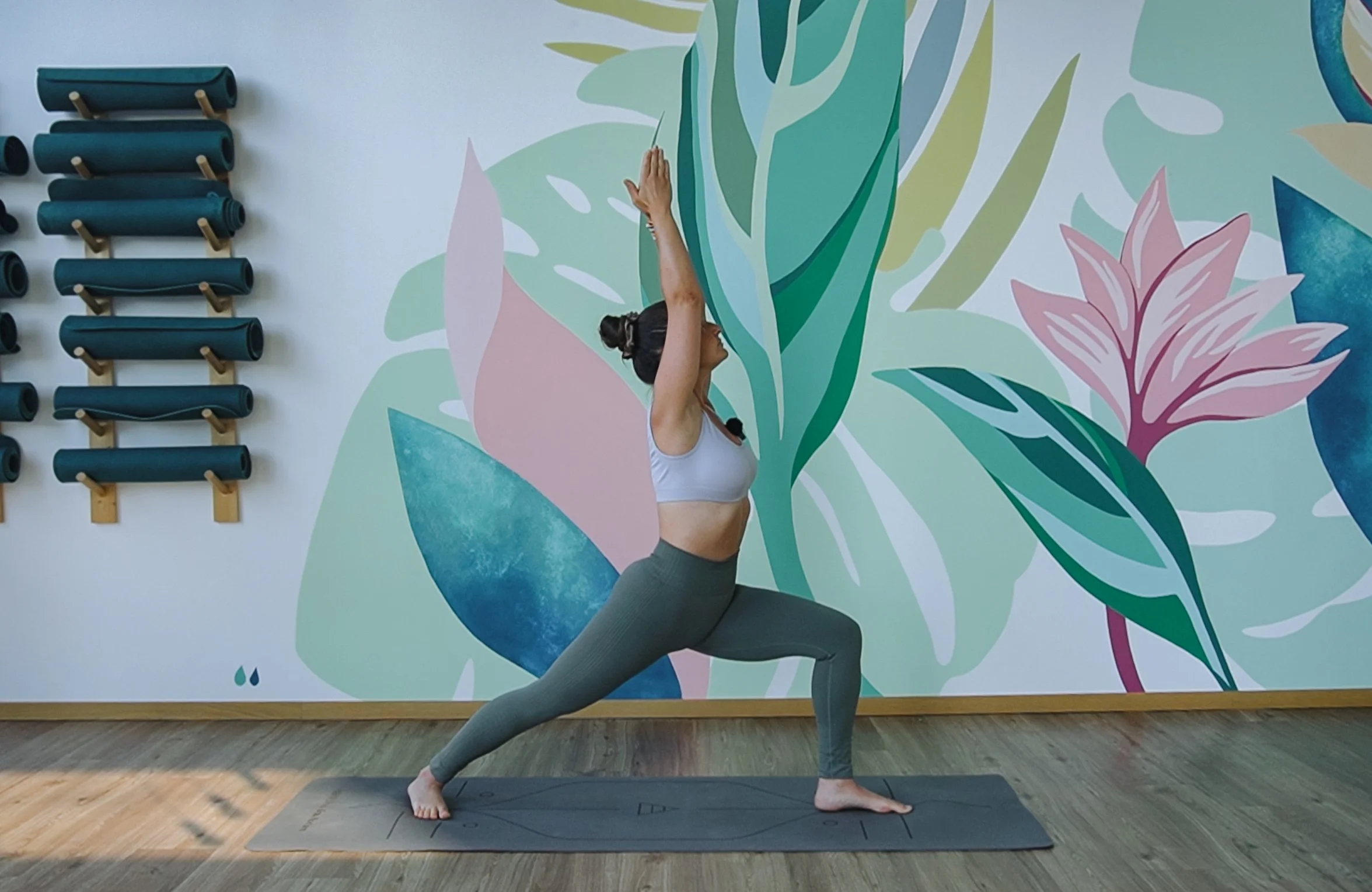 Femme faisant du yoga en position de guerrier dans une salle avec un mur peint de motifs floraux et feuilles