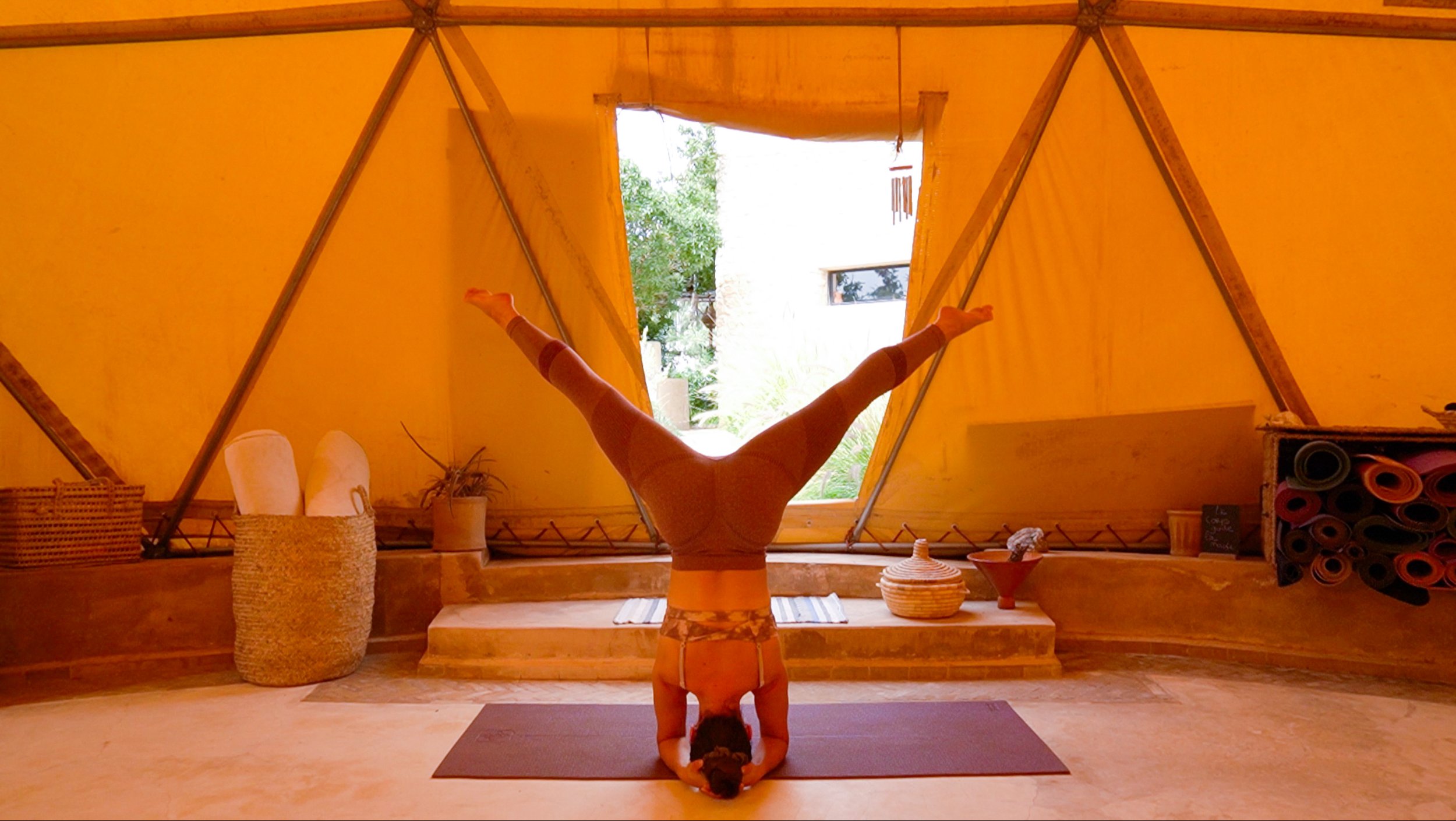 Une personne pratiquant le yoga en position de tête, jambes écartées, dans une tente en bois avec vue sur la nature extérieure.