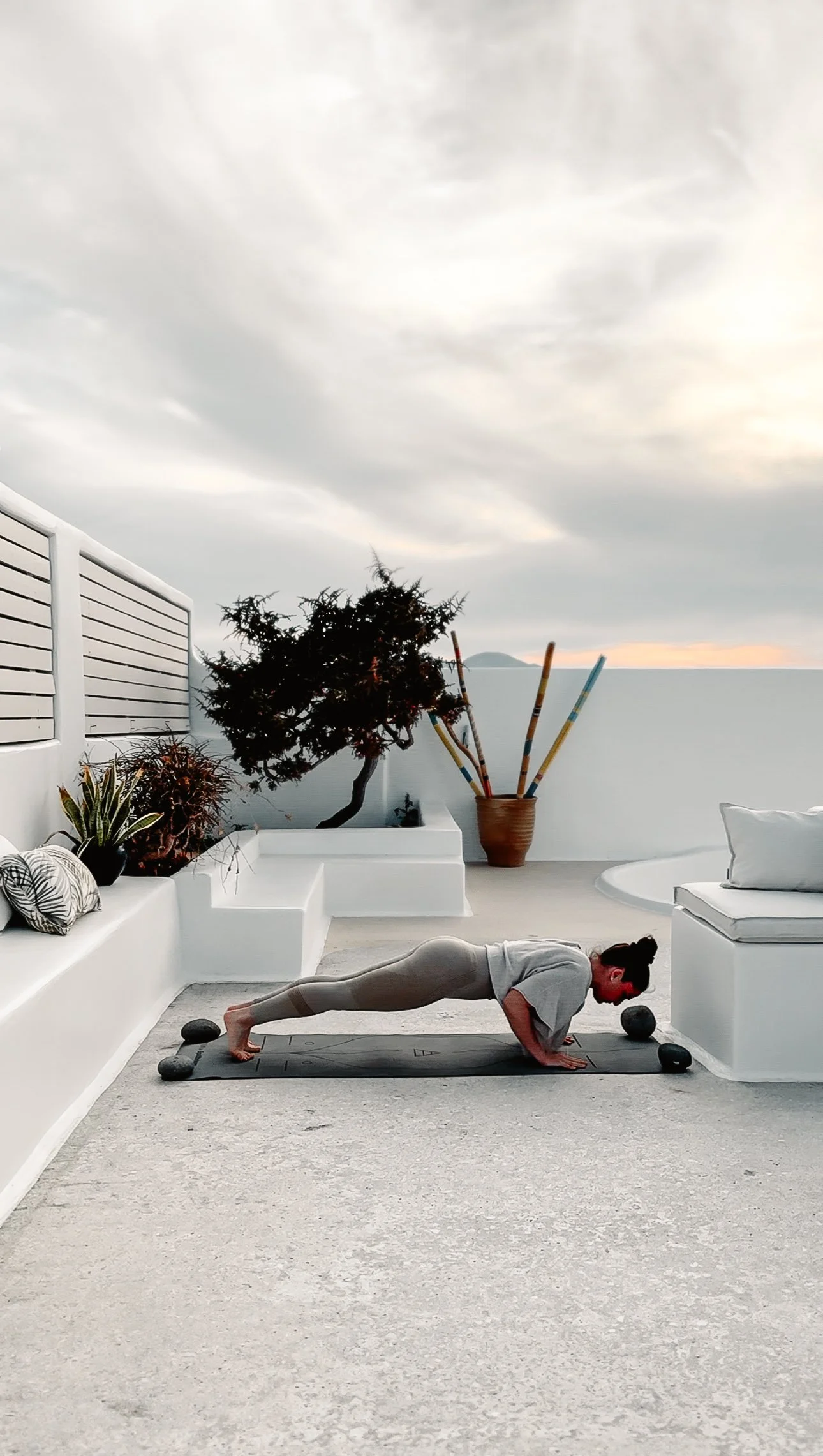 Une femme pratique une position de planche sur un tapis de yoga en extérieur, décoré avec des plantes et un ciel nuageux en arrière-plan.