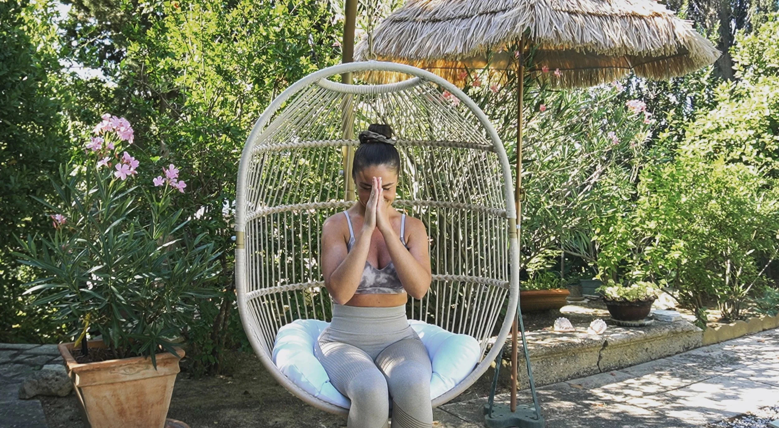 Femme pratiquant la méditation en position de prière dans un jardin avec un fauteuil suspendu en rotin, entourée de plantes et de fleurs.