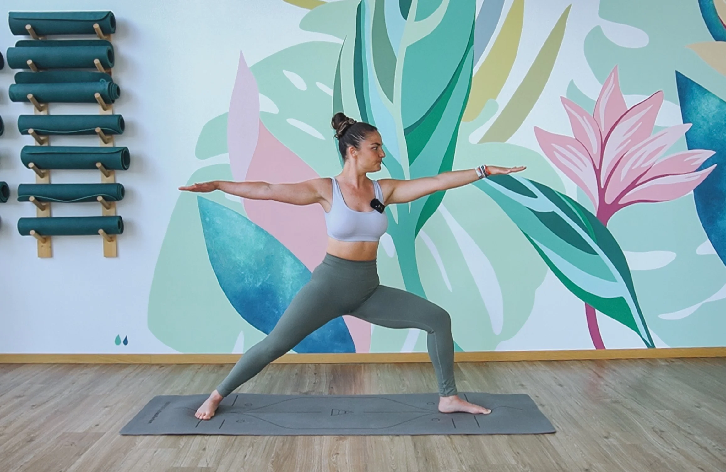 Femme pratiquant le yoga en position de guerrier dans une salle de sport avec mur décoré de fleurs et plante stylisées