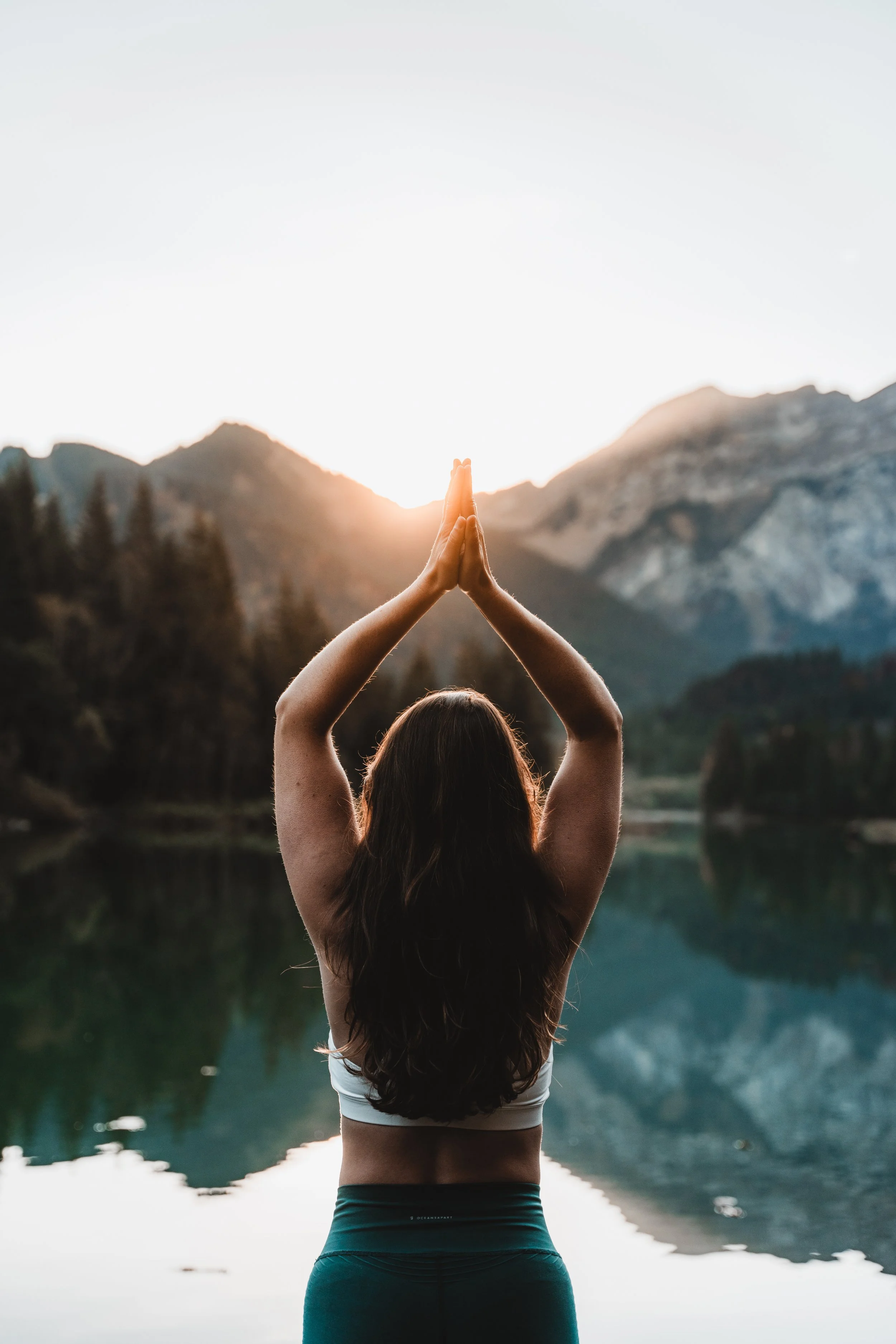 Femme pratiquant la méditation ou le yoga au bord d'un lac, avec des montagnes en arrière-plan, au lever ou coucher du soleil.