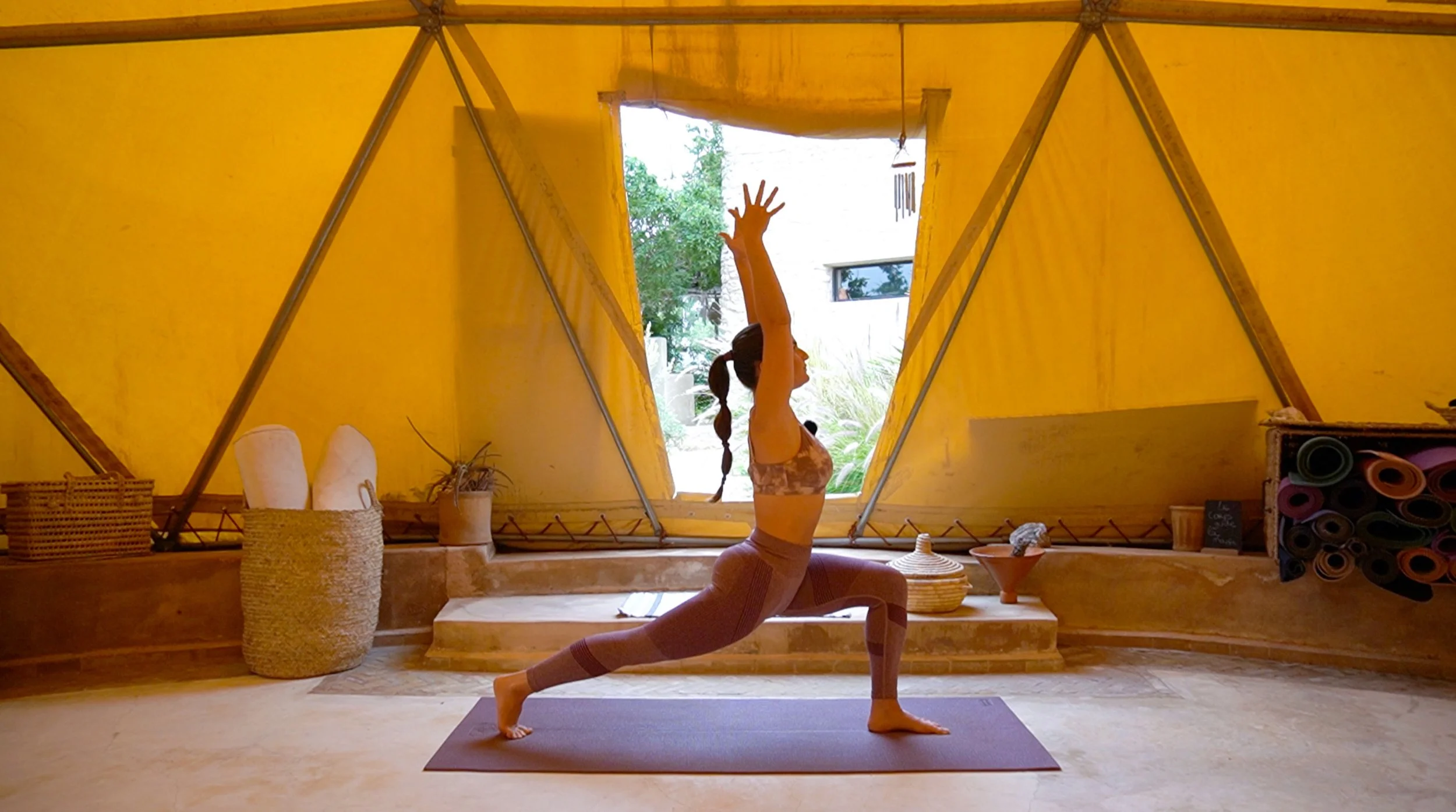 Femme faisant du yoga dans un chalet lumineux avec murs jaunes, pratiquant une posture de guerrier, tapis de yoga, décor naturel avec plantes et objets de décoration