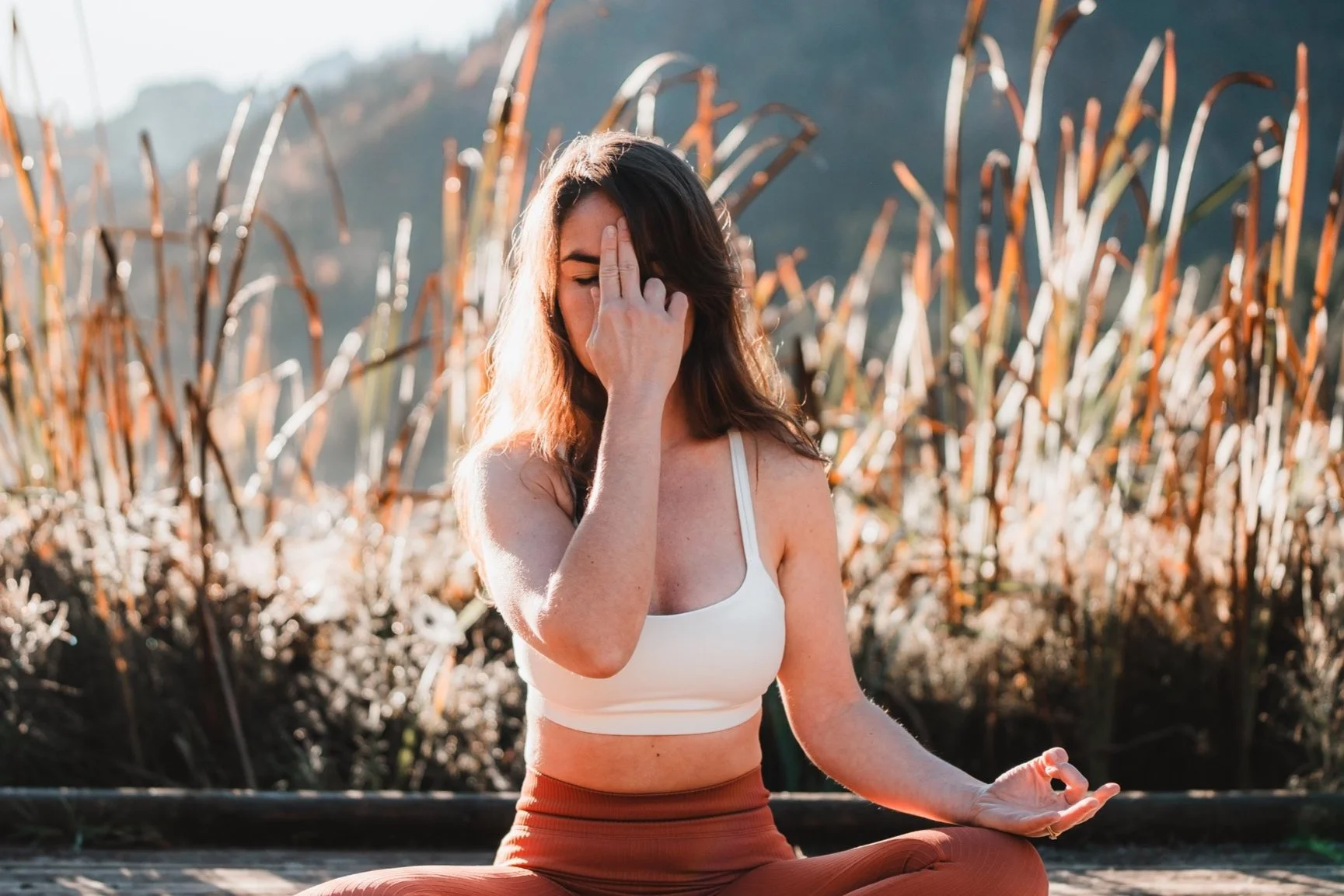 Femme méditant en position de yoga en plein air, entourée de plantes sèches, probablement dans un champ ou en nature, lors d'une séance de bien-être.