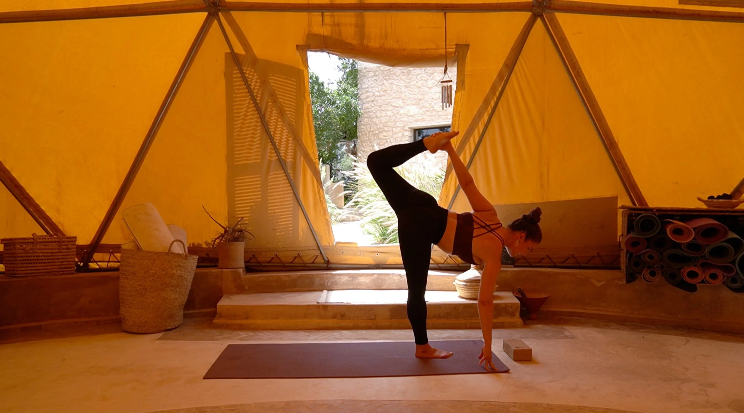 Femme pratiquant le yoga dans une tente en tissu jaune, faisant une posture de yoga avec un bras et une jambe levés, un tapis de yoga et des rouleaux de tapis en arrière-plan.