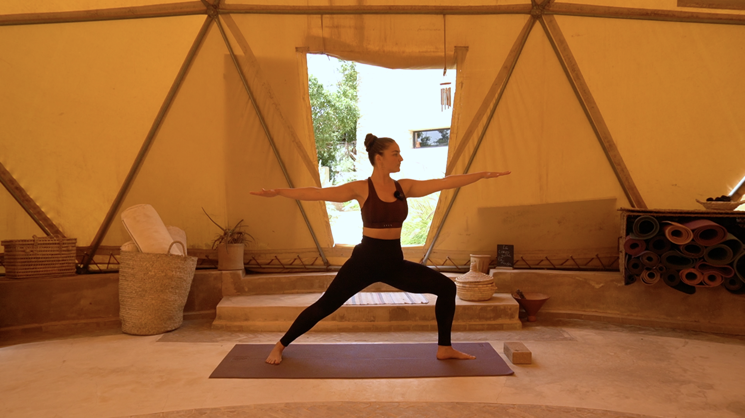 Une femme pratique le yoga en position de guerrier dans une tente jaune avec des rouleaux de tapis de yoga empilés à droite.