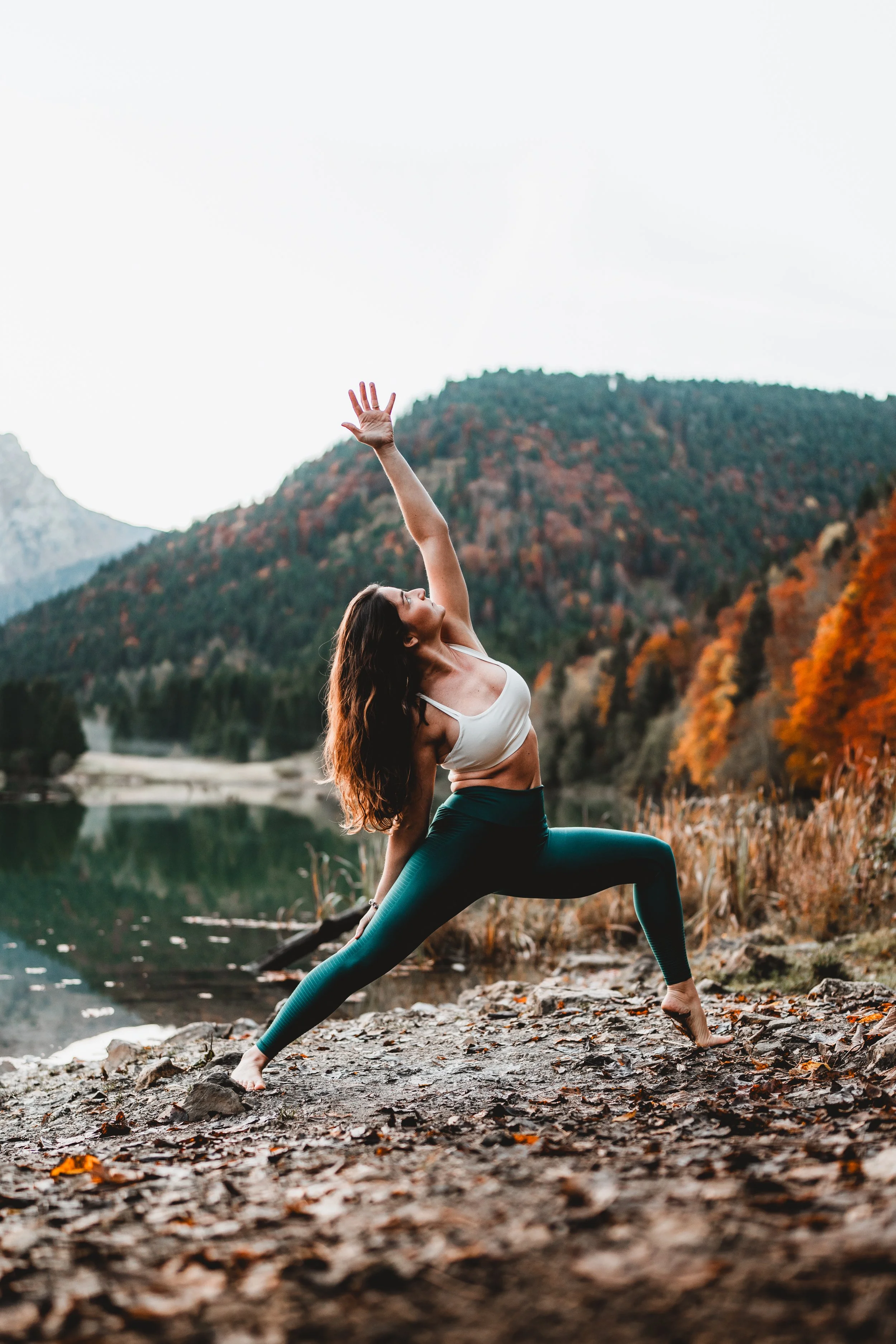 Femme pratiquant le yoga en extérieur sur un terrain rocheux près d'un lac, avec des montagnes et des arbres aux feuillages d'automne en arrière-plan.