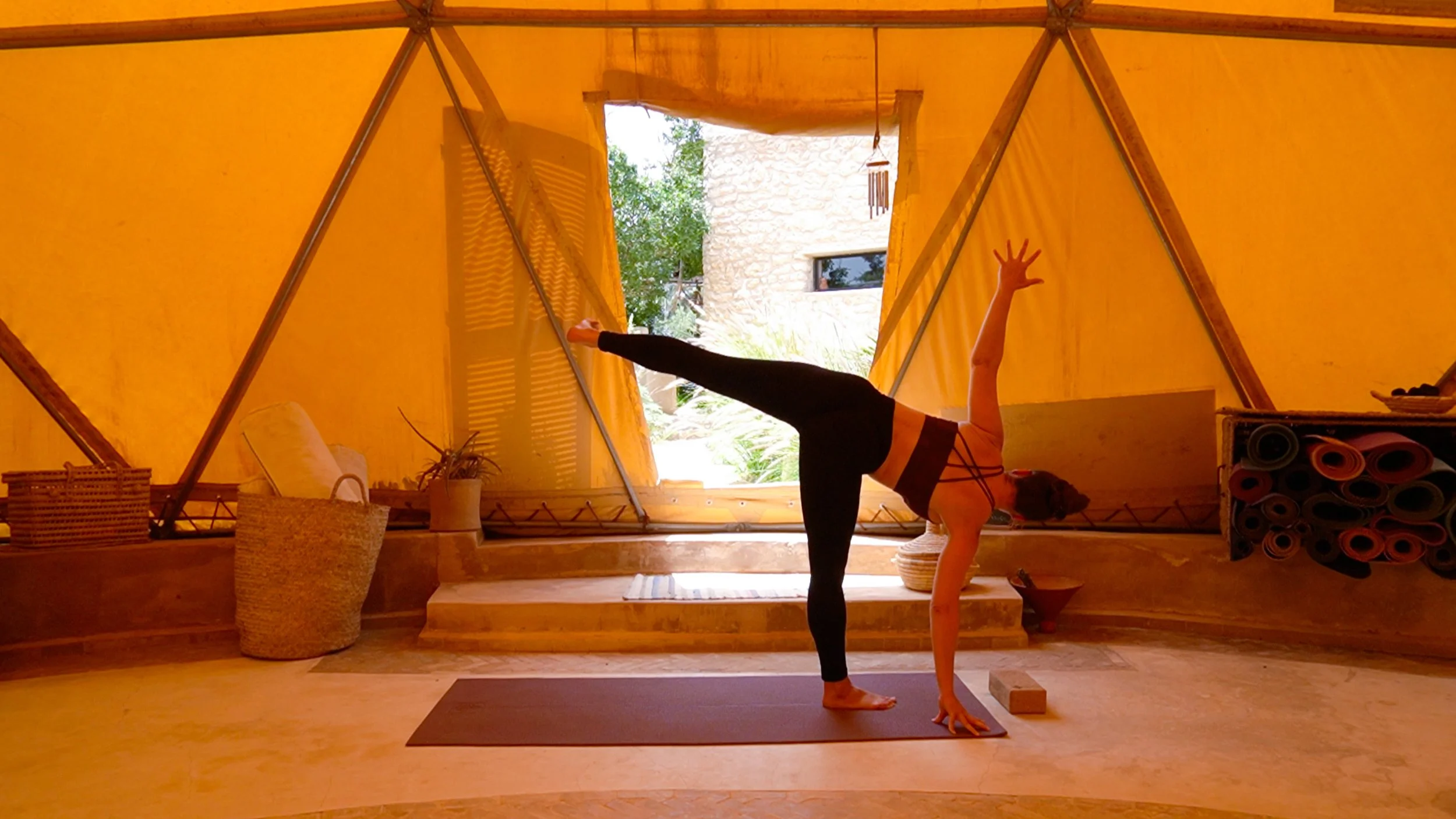 Femme faisant du yoga dans une tente en tissu orange avec vue sur le jardin, portant un haut noir et leggings noir, en position du chien tête en bas.