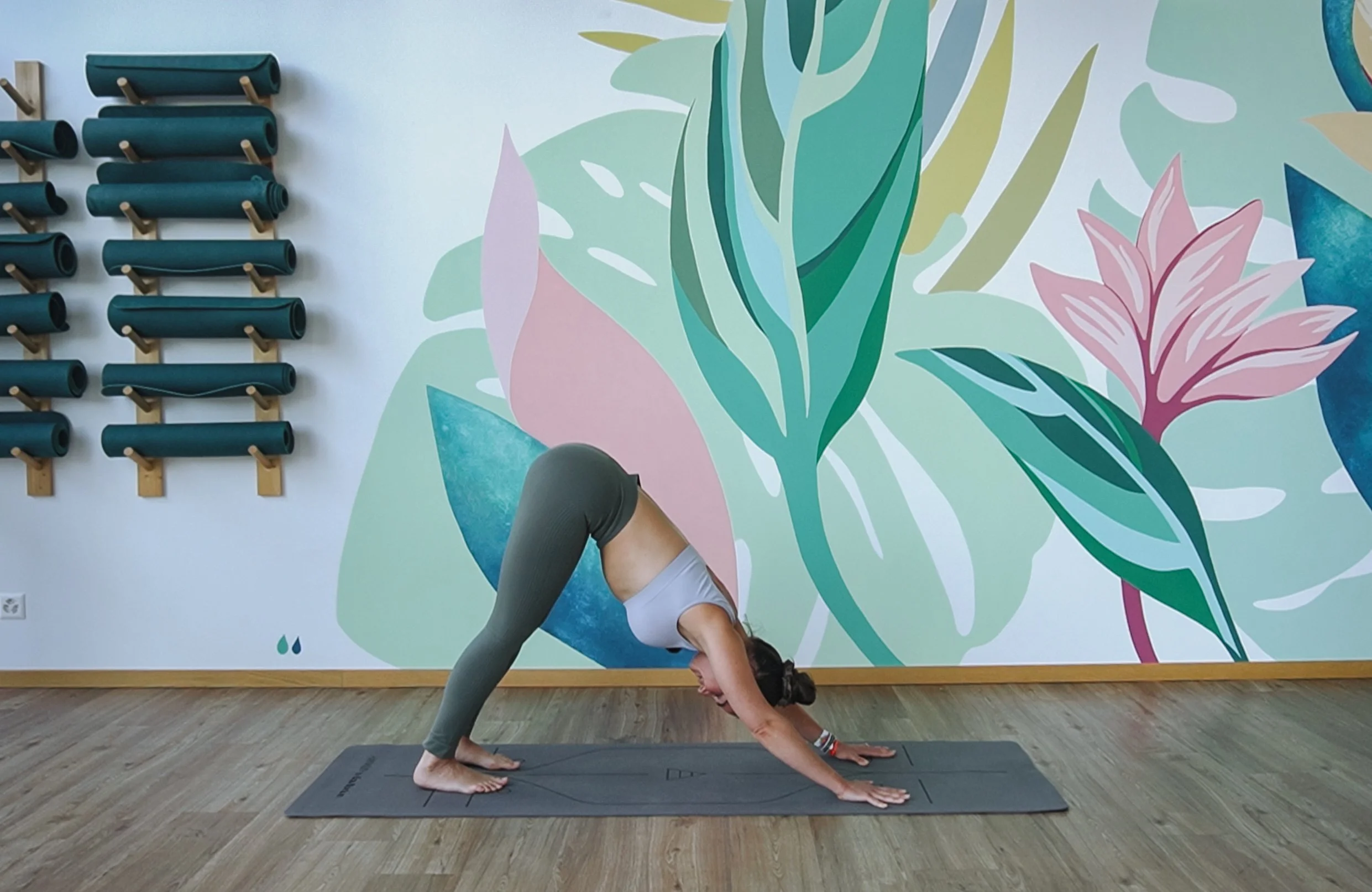 Une femme pratique le yoga dans une salle avec un mur décoratif floral, en position de chien tête en bas sur un tapis de yoga.