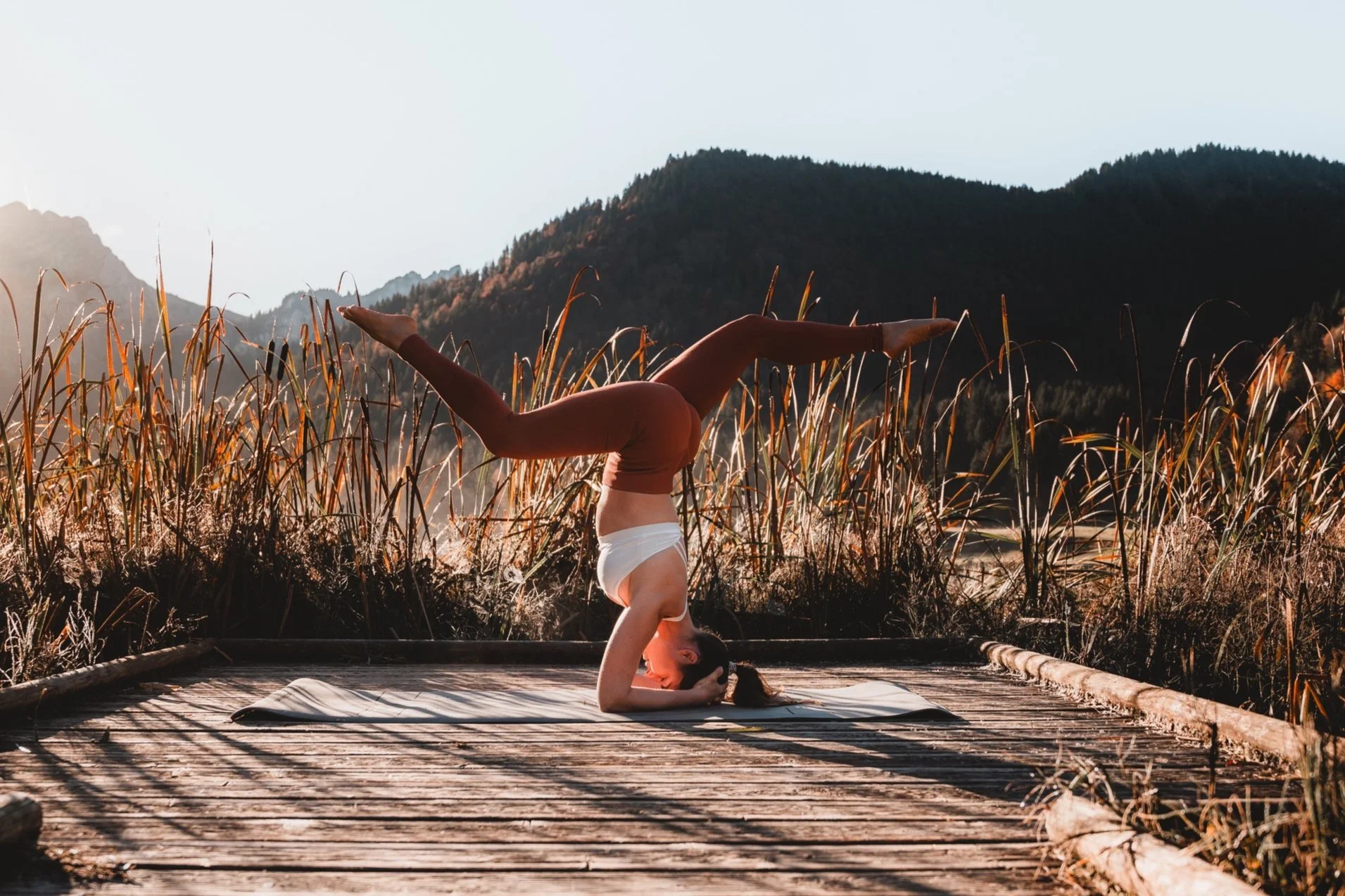 Femme pratiquant la respiration de yoga sur un tapis en bois au coucher du soleil dans un paysage de montagnes et de hautes herbes.