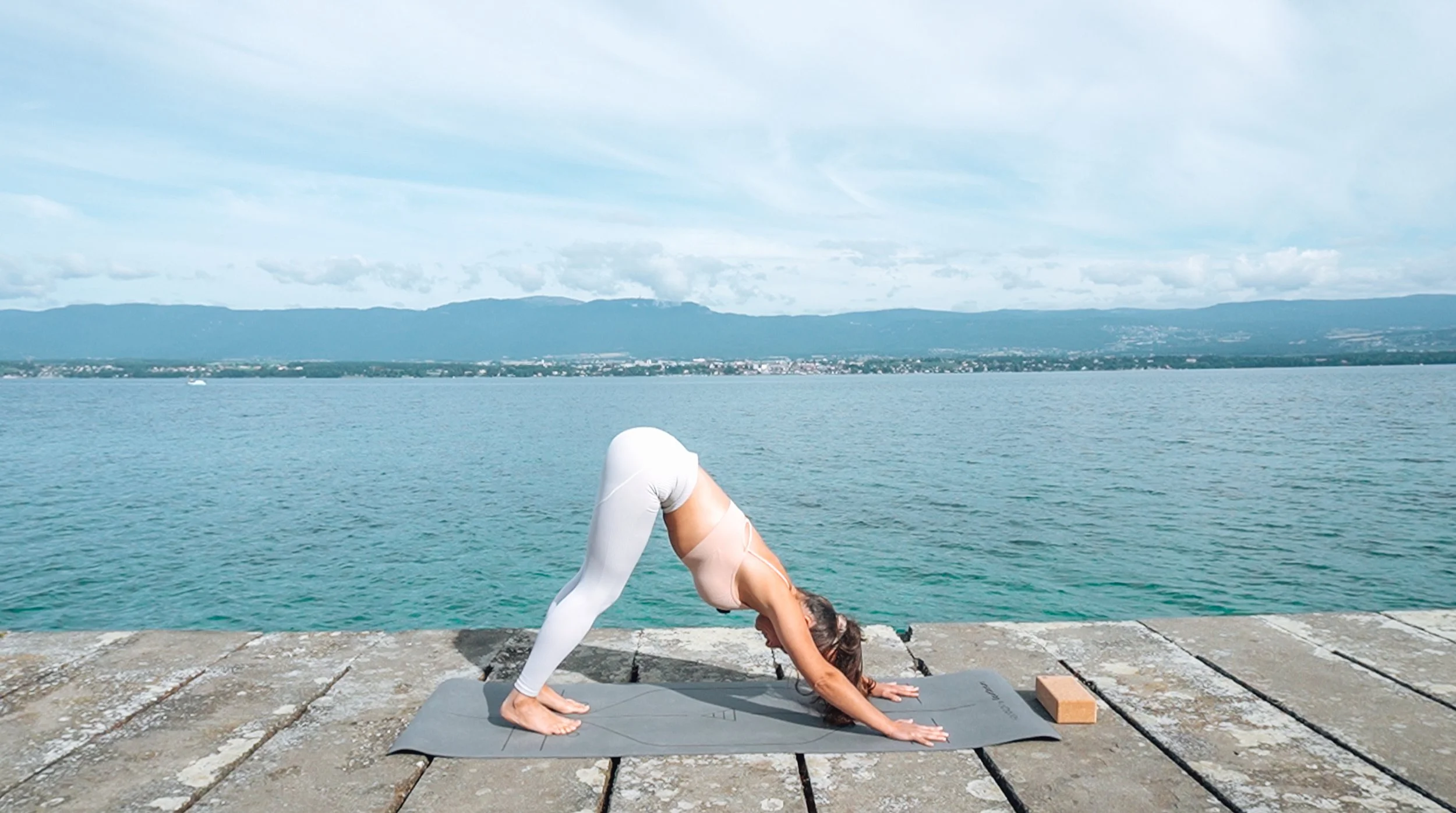 Femme pratiquant le yoga en posture du chien tête en bas sur une plateforme en pierre au bord de l'eau avec un ciel partiellement nuageux et des montagnes à l'arrière-plan.