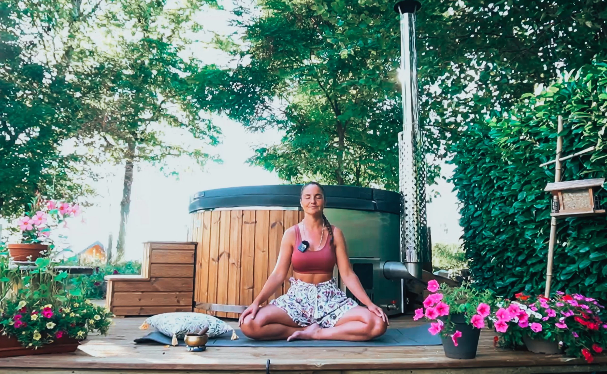 Femme faisant de la méditation en plein air, assise en position du lotus sur une plateforme en bois, entourée de fleurs et de verdure, avec un bain à remous en arrière-plan.