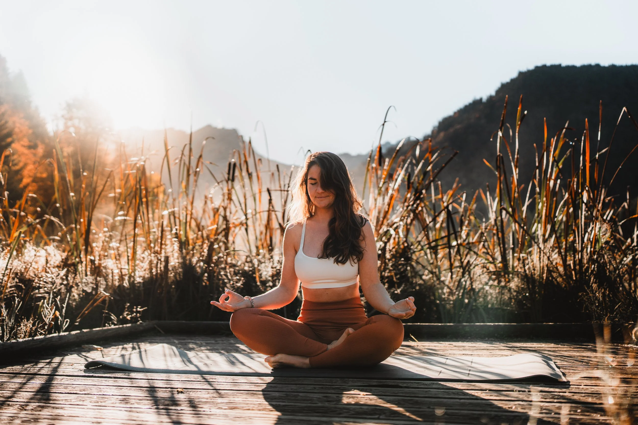 Femme méditant en position de lotus sur un tapis de yoga en plein air avec un paysage de marais et de montagnes au coucher du soleil.