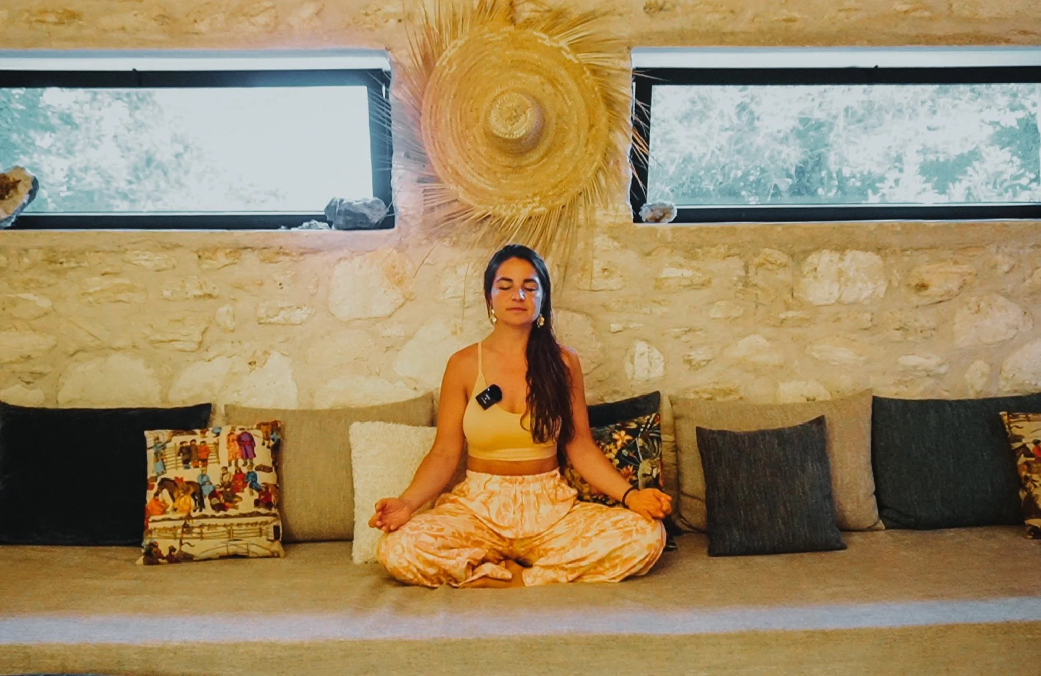 Femme méditant en position de lotus sur un canapé avec des coussins, dans une pièce à mur en pierre et deux fenêtres rectangulaires, décorée d'un grand motif en osier au mur.