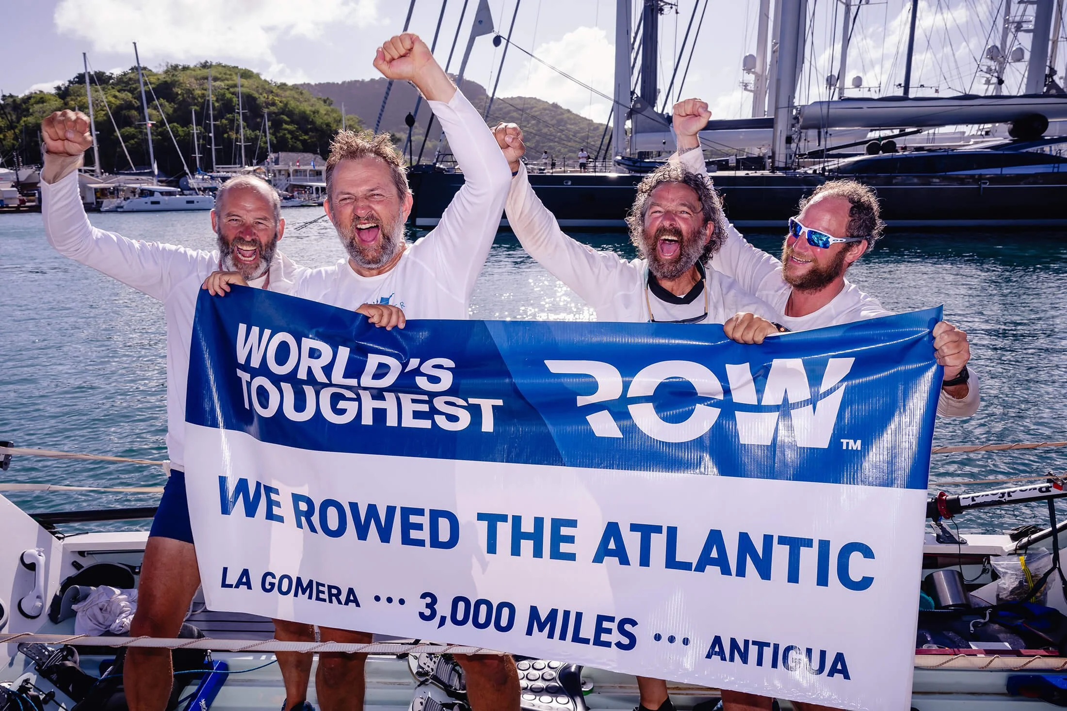 Four men celebrating on a boat, holding a banner reading 'World's Toughest Row, We Rowed the Atlantic, La Gomera 3,000 Miles, Antigua.' They are smiling, with some raising their fists in victory, and are in a marina with sailboats and lush green hill