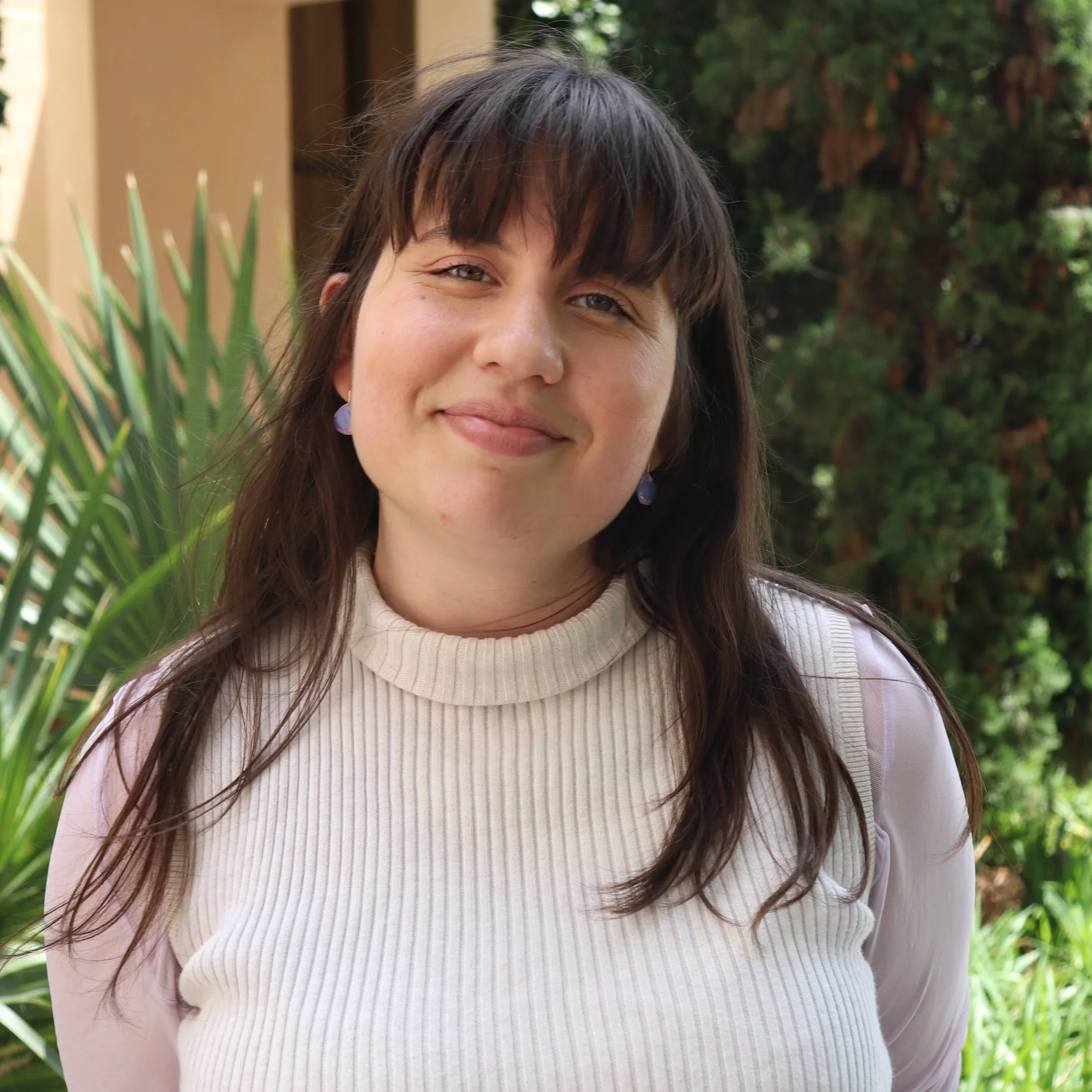 A woman with dark brown hair and bangs, smiling outdoors with green plants and trees in the background, wearing a light cream-colored ribbed sweater and purple earrings.
