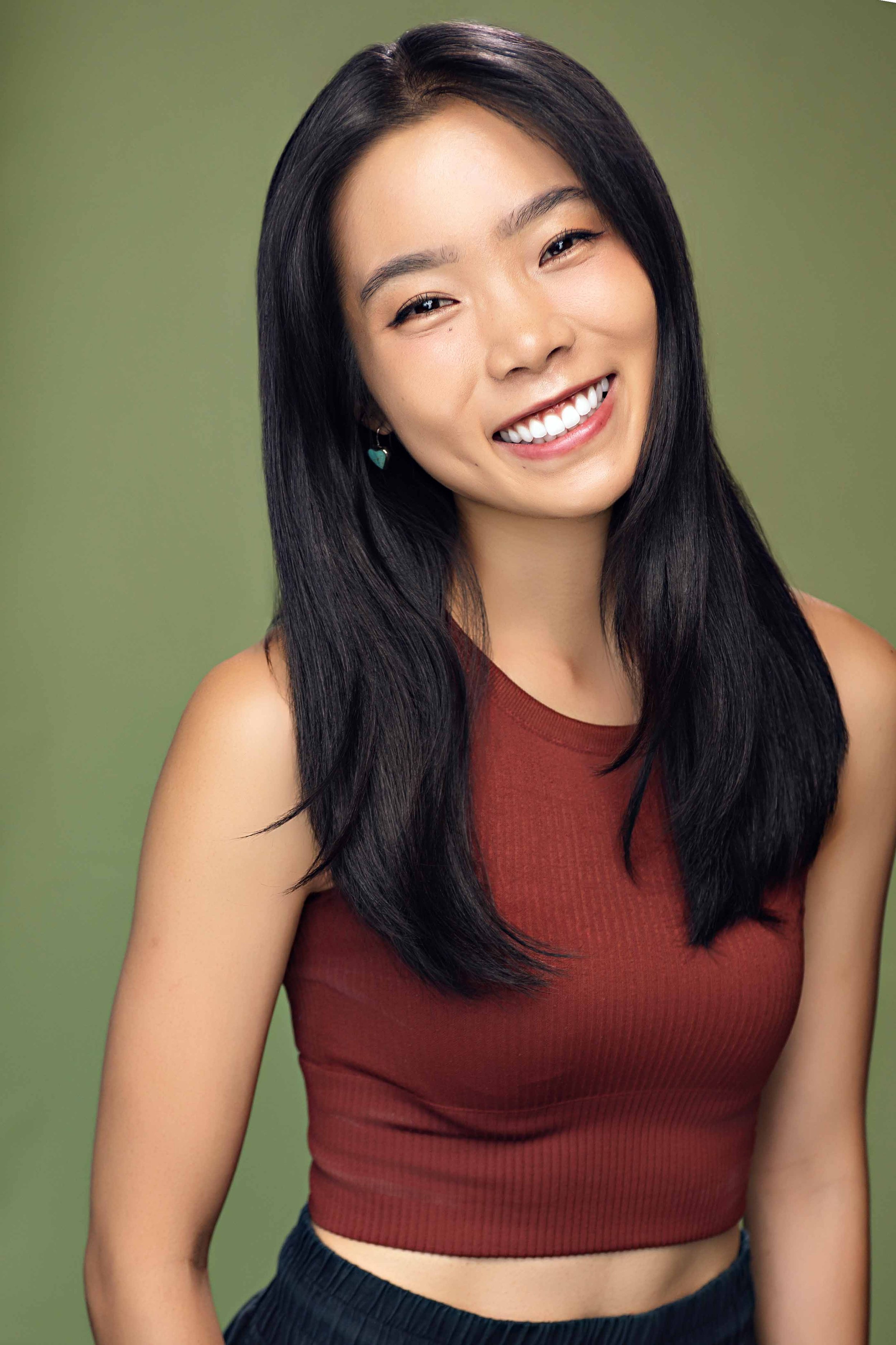 A young woman with long black hair smiling, wearing a sleeveless maroon top and earrings, against a green background.