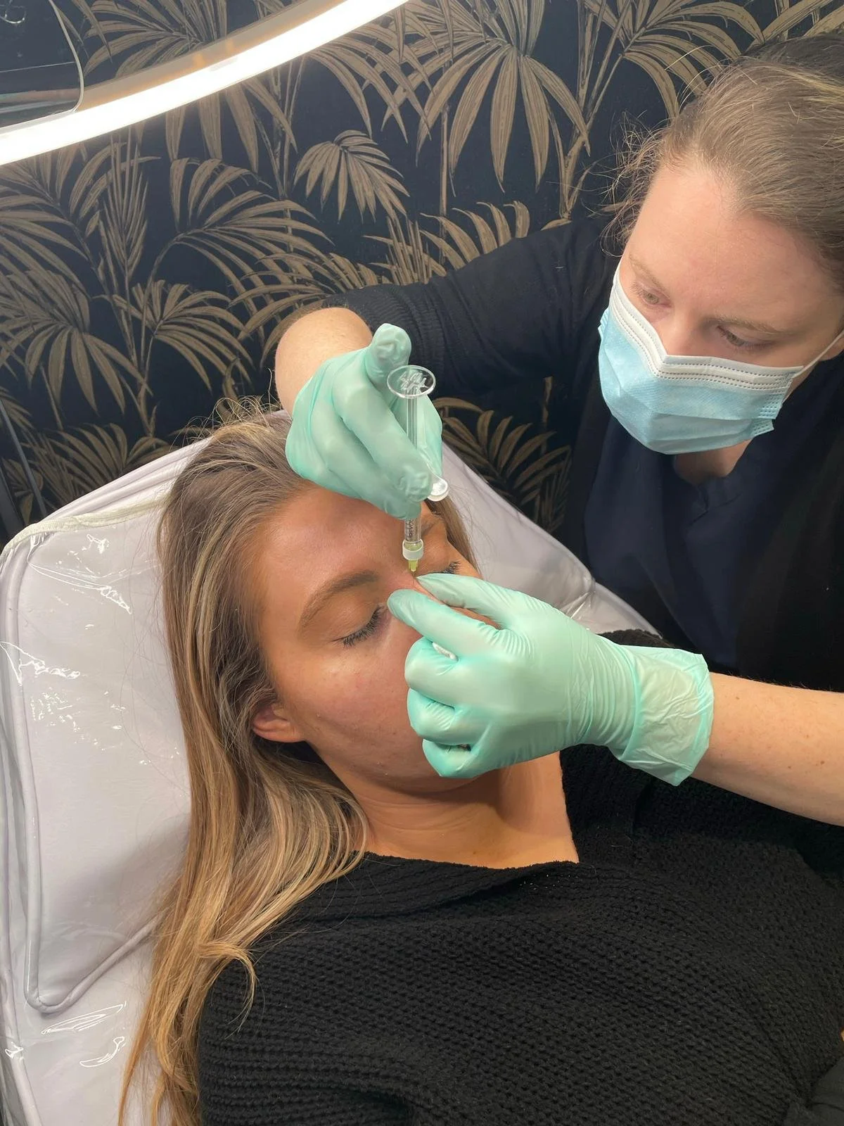 A woman lying on a treatment bed receiving an injection in her forehead from a healthcare professional wearing gloves and a face mask, with a patterned wall in the background.