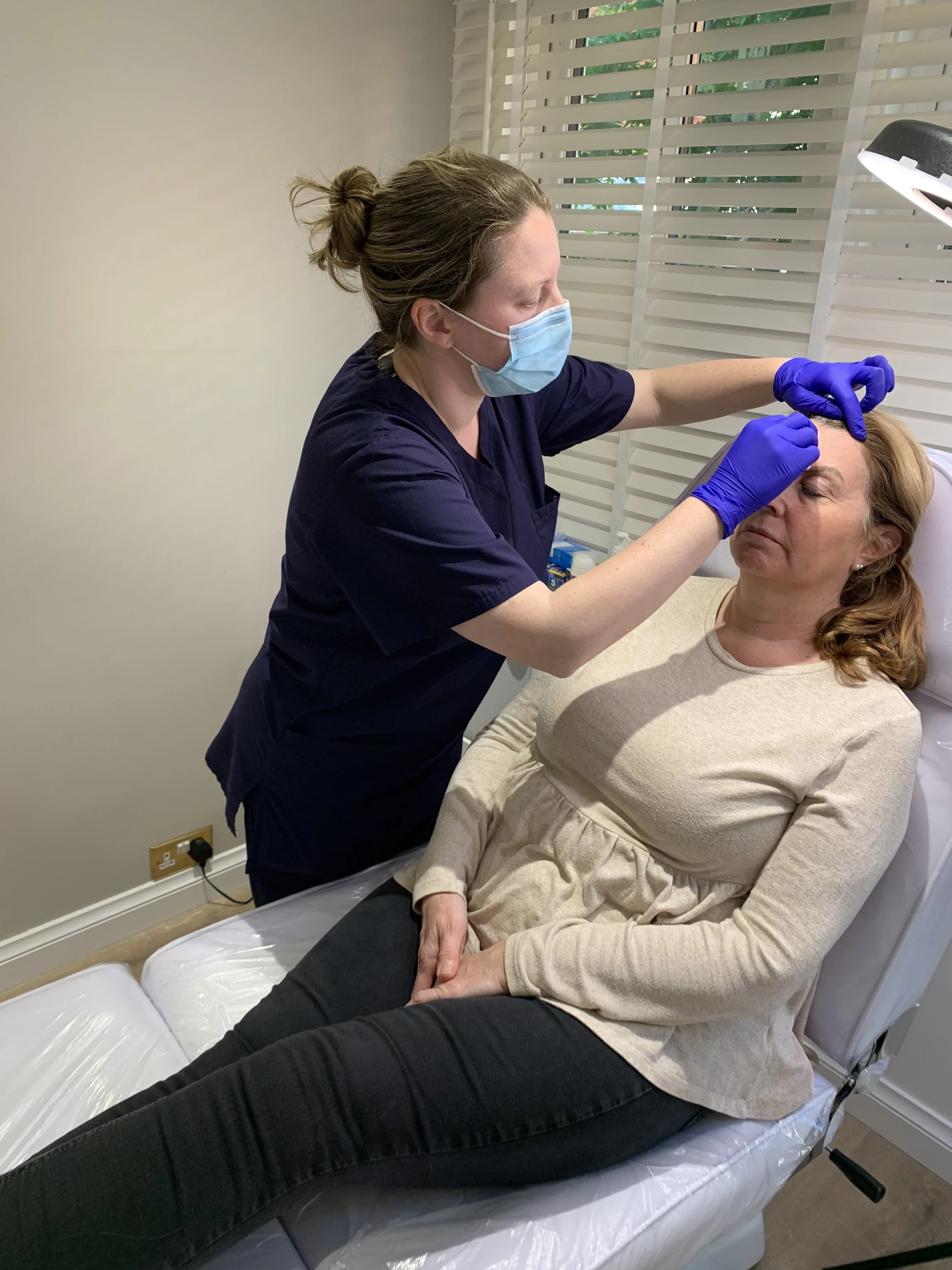 A healthcare professional wearing a mask and gloves administering a cosmetic or medical procedure to a woman's face in a clinical setting.