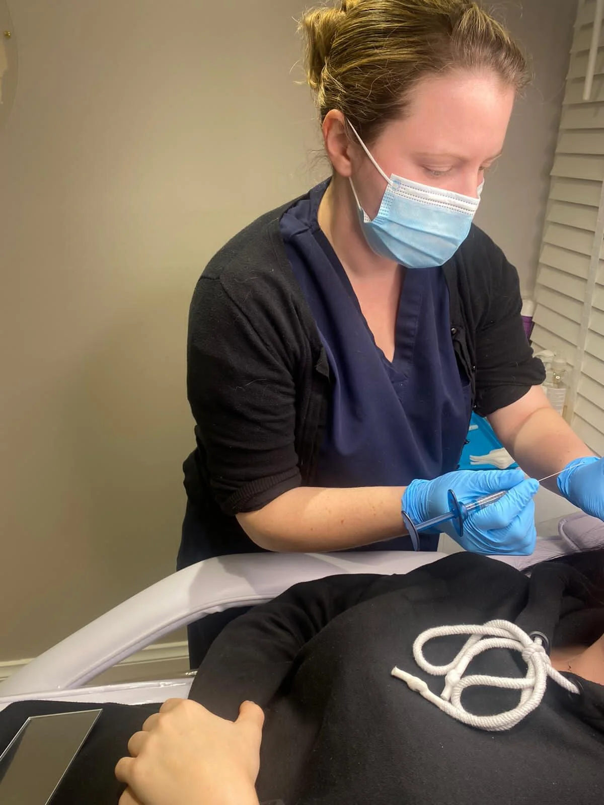 A healthcare professional in scrubs, wearing a face mask and gloves, preparing to give an injection to a patient lying on a medical bed.