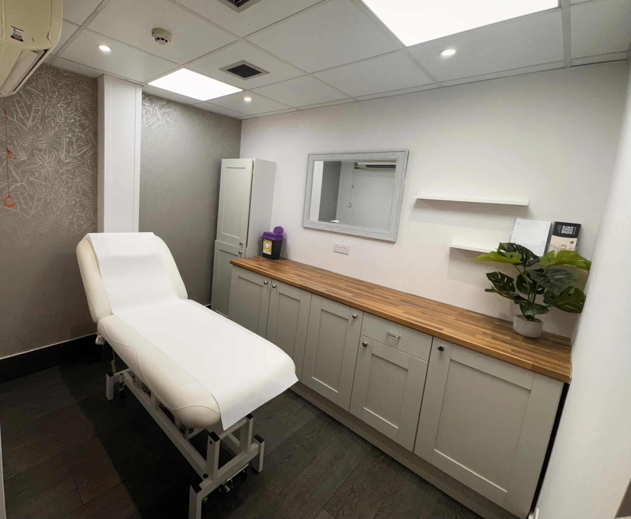 A medical examination room showing an examination bed, white cabinetry, a mirror on the wall, and some potted plants, with a wood countertop and patterned wall wallpaper.