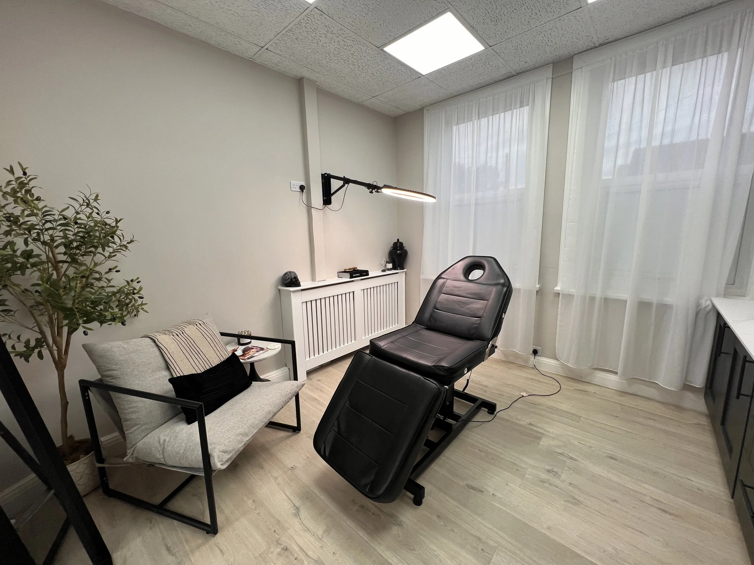 A consultation or therapy room with two chairs, a small white side table, white radiator cover, potted plant, wall-mounted light, books, and a large window with sheer curtains.
