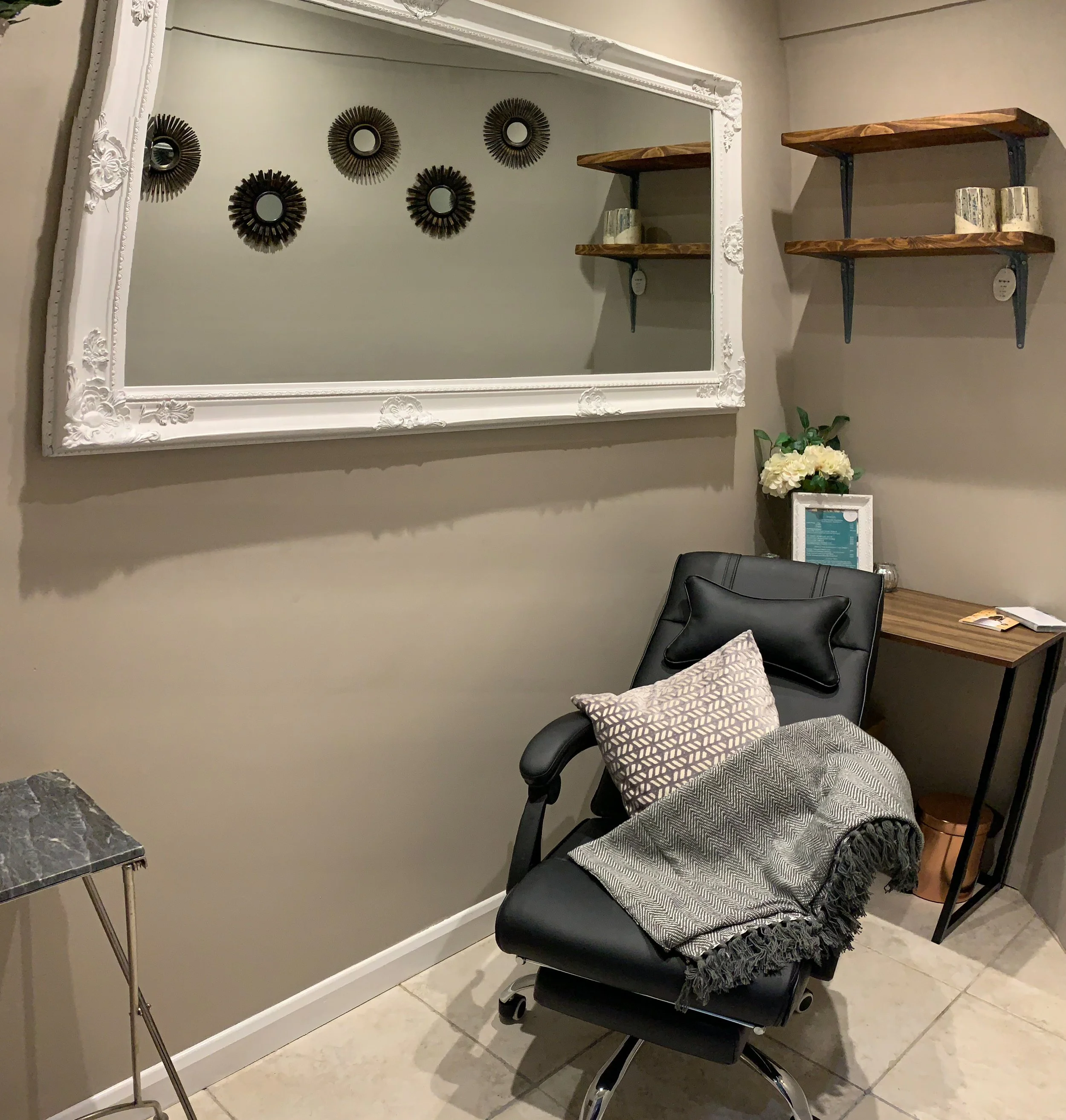 A black office chair with a cushioned seat and backrest, a patterned pillow, and a gray throw blanket on it. Behind the chair, there are wooden and metal wall shelves with candles. To the left, part of a marble-topped table is visible. On the wall, there is a large ornate framed mirror and five decorative wall mirrors with sunburst designs. A small potted plant with white flowers is on a table next to the chair.