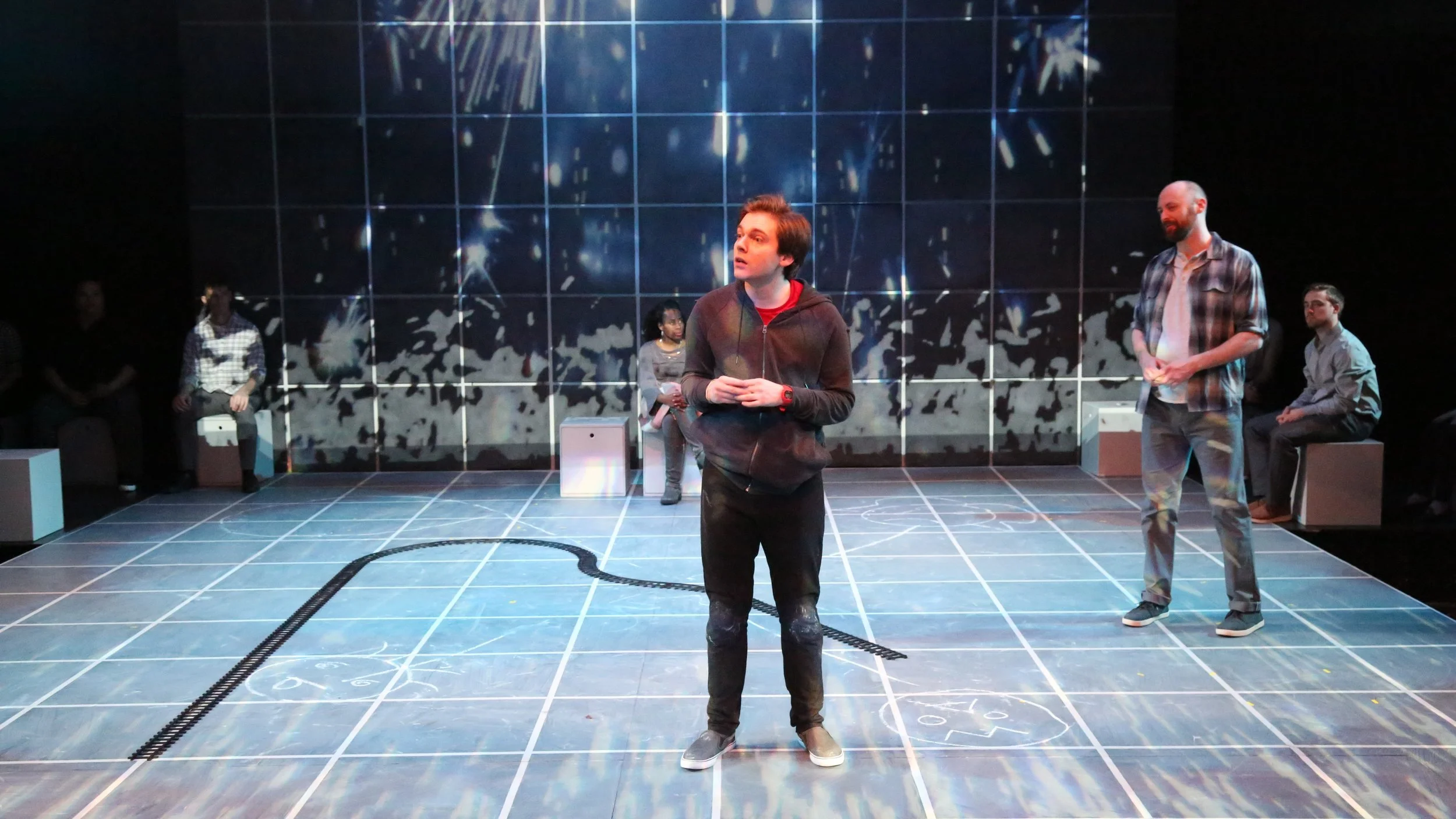 Performance image from The Curious Incident of the Dog at Night Time. Chris is standing in a group of people staring at him. Red spots are projected all over the envrionment.