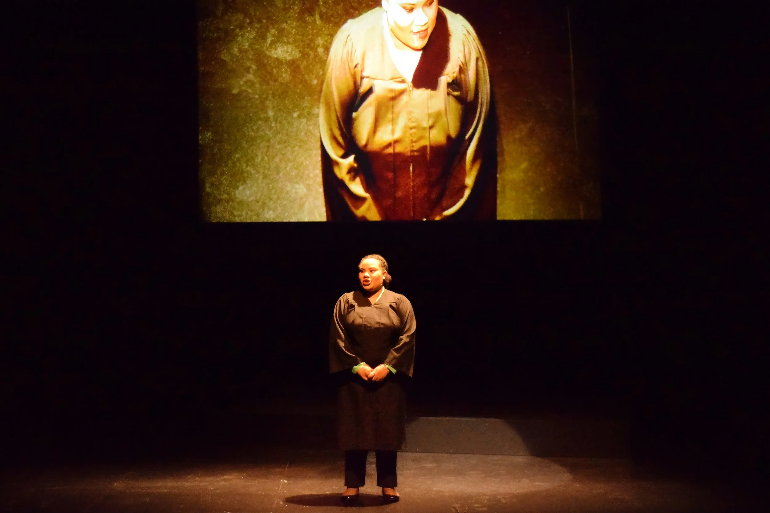 Performance image from Execution of Justice. A judge stands before a large screen showing a top down image the judge from a security camera.