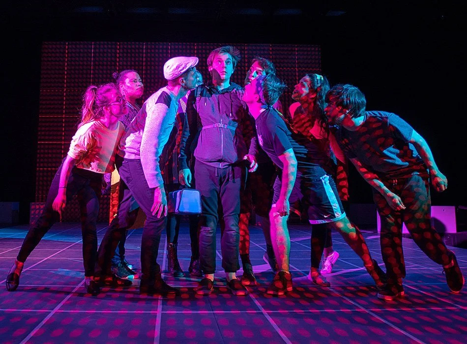 Performance image from The Curious Incident of the Dog at Night Time. Chris is standing in a group of people staring at him. Red spots are projected all over the envrionment.