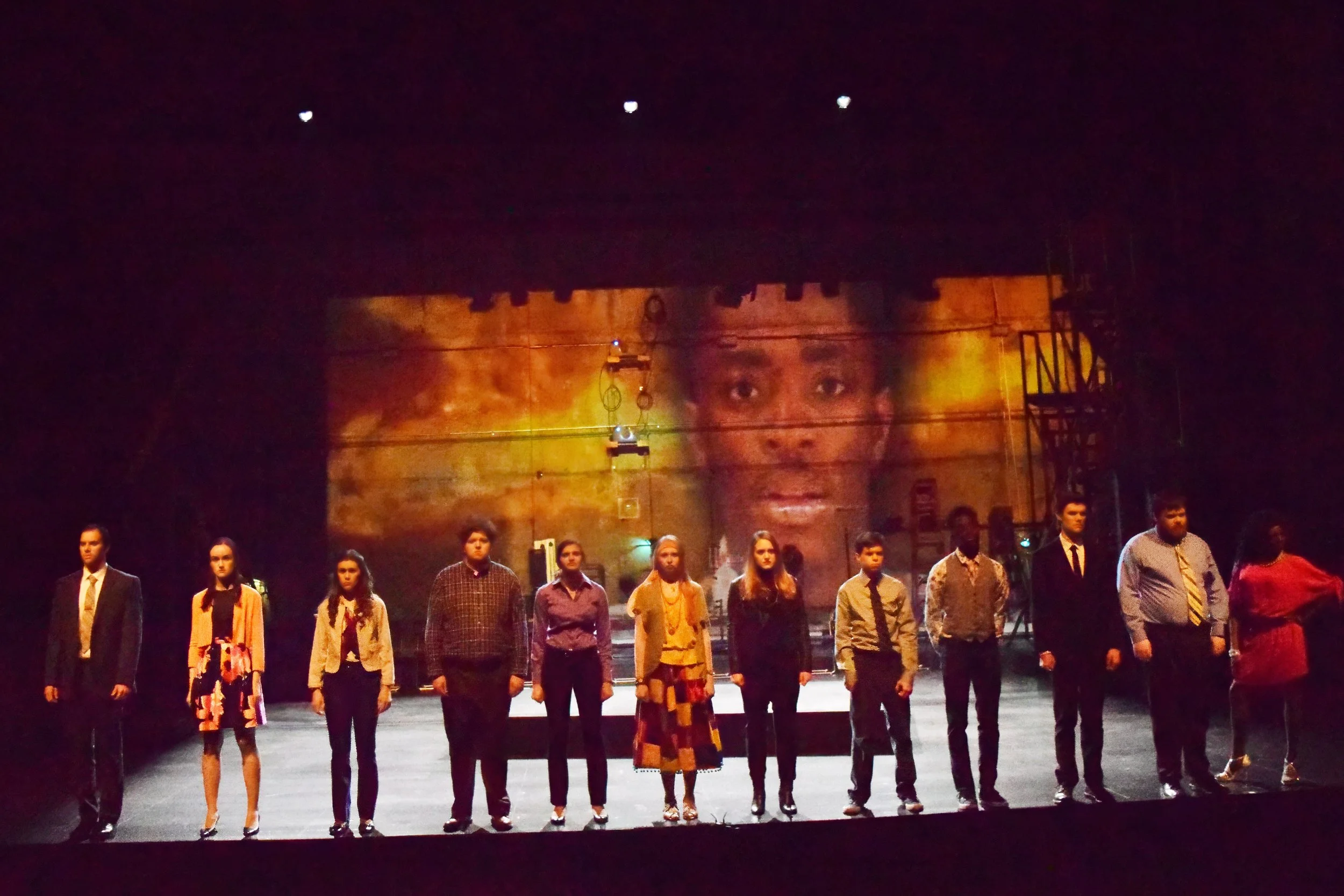 Execution of Justice production photo. Cast members stand at the edge of the stage apron in front of a large black wall a projection of a young black man talking in front of a field of fire.