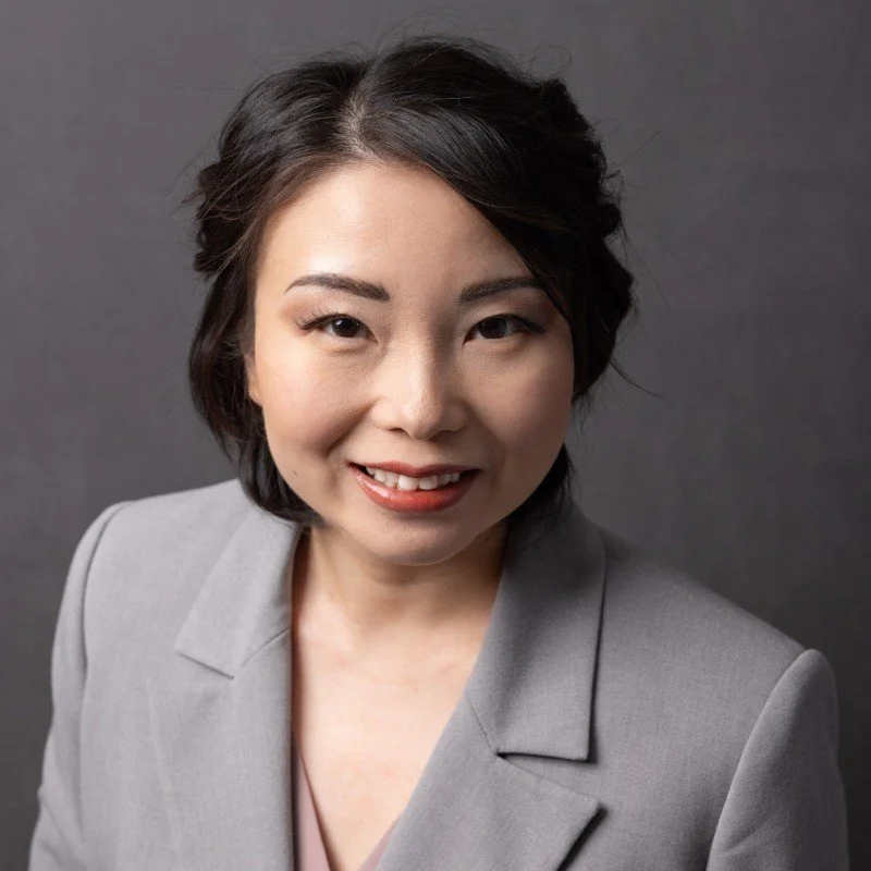A woman with short black hair smiling, wearing a light gray blazer, against a dark background. Shanshan Thompson