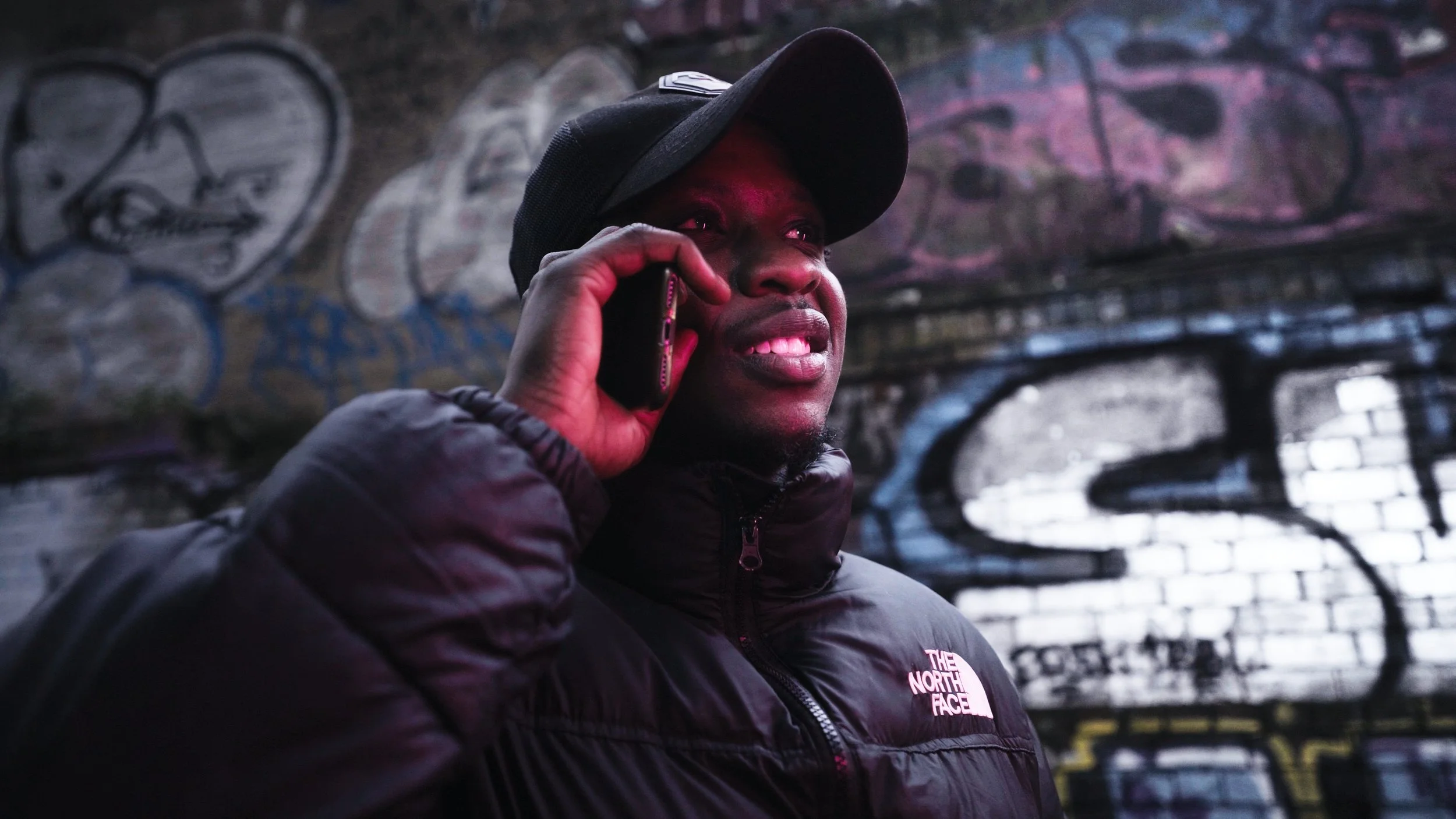 Person in black jacket and cap talking on a phone with graffiti-covered wall in the background.